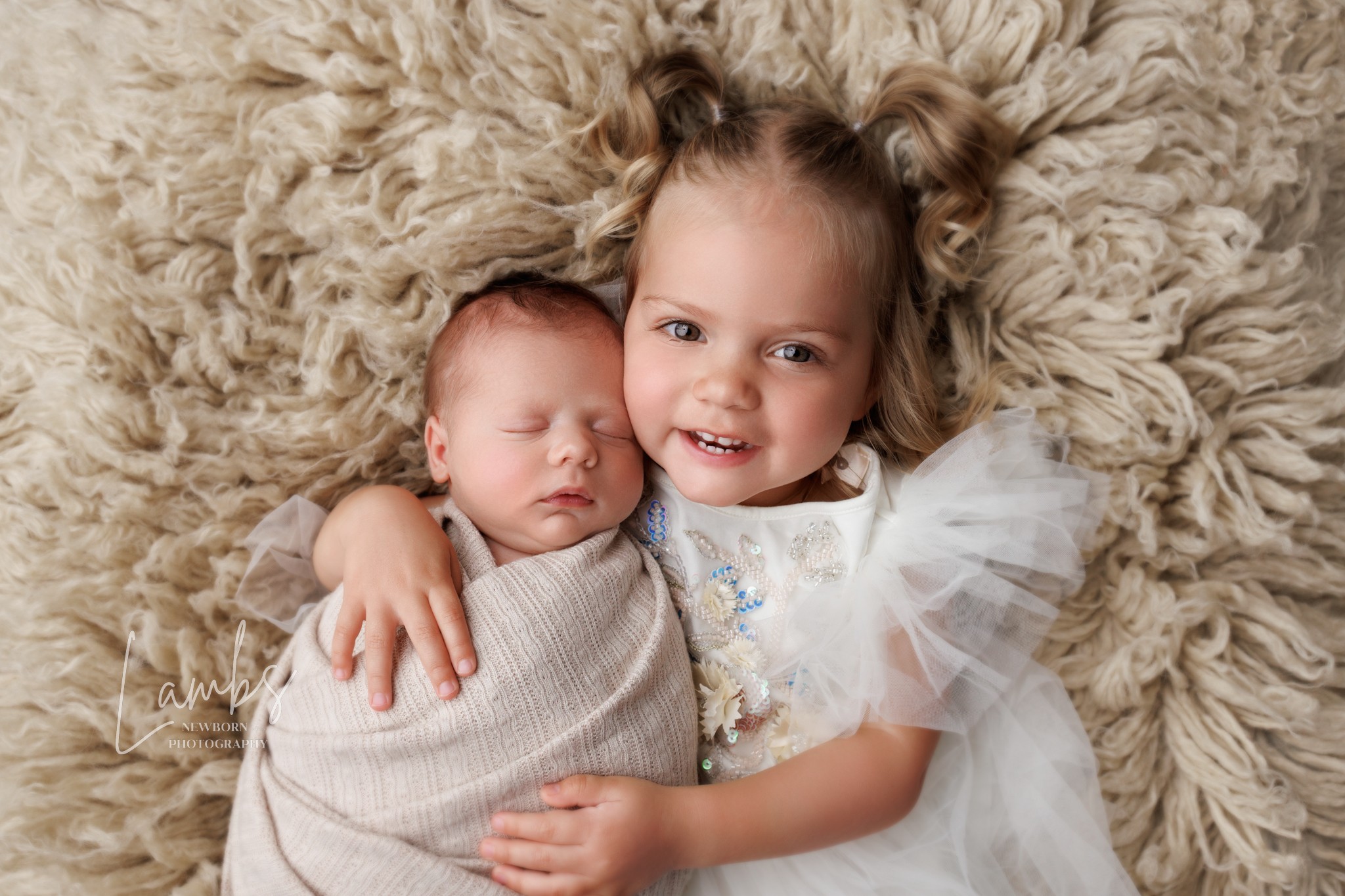 Two young children, one a newborn and the other a toddler, lying on a fluffy beige rug, smiling and hugging.