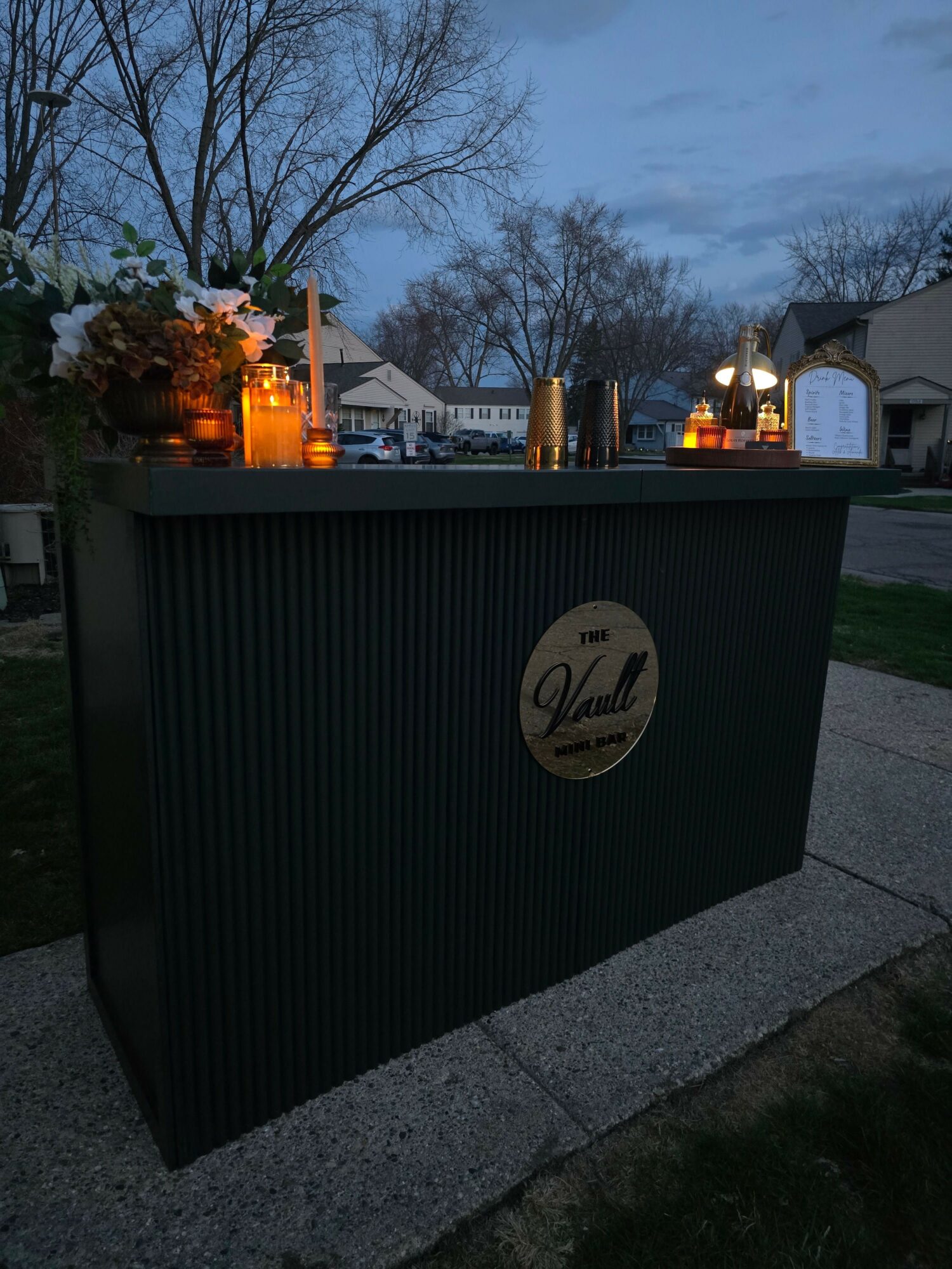Black sign with a circular logo and text, trees, and buildings in the background during evening.