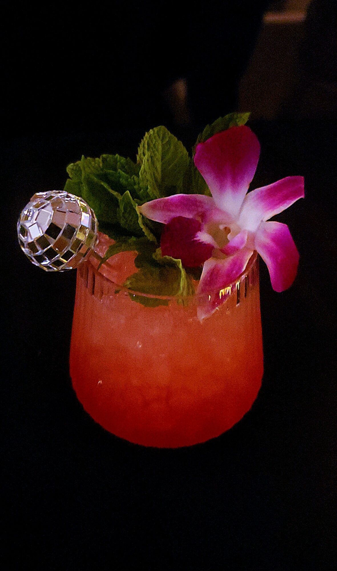 Colorful cocktail in a glass with a pink flower, green leaves, and a small disco ball decoration, against a dark background.