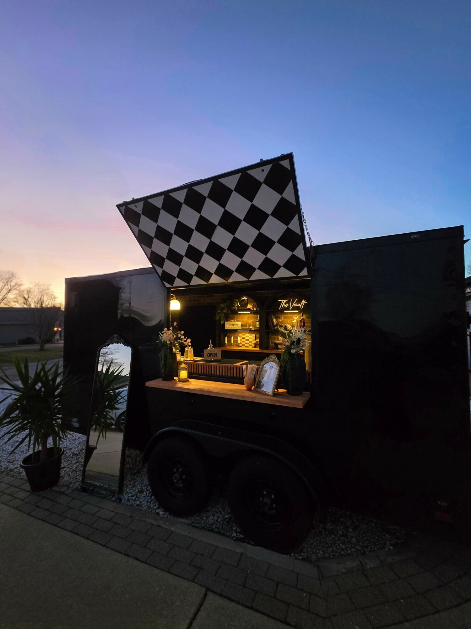 A black mobile bar with a checkered canopy, set outdoors during sunset, with plants nearby.