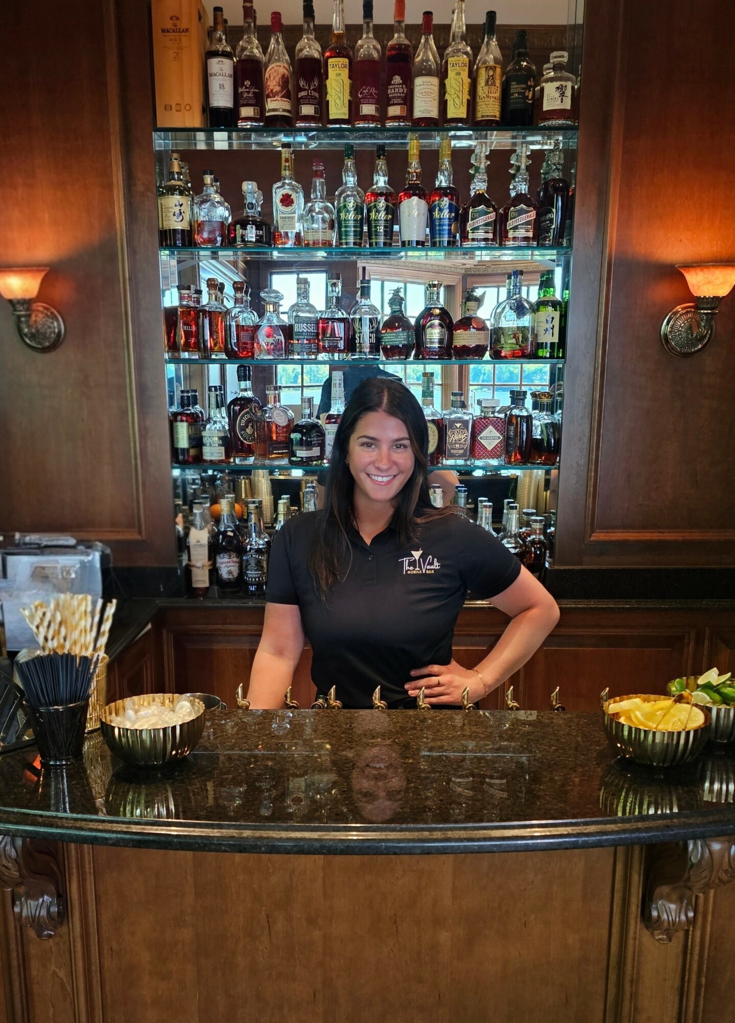 Smiling woman standing behind a bar with shelves of liquor bottles in the background.