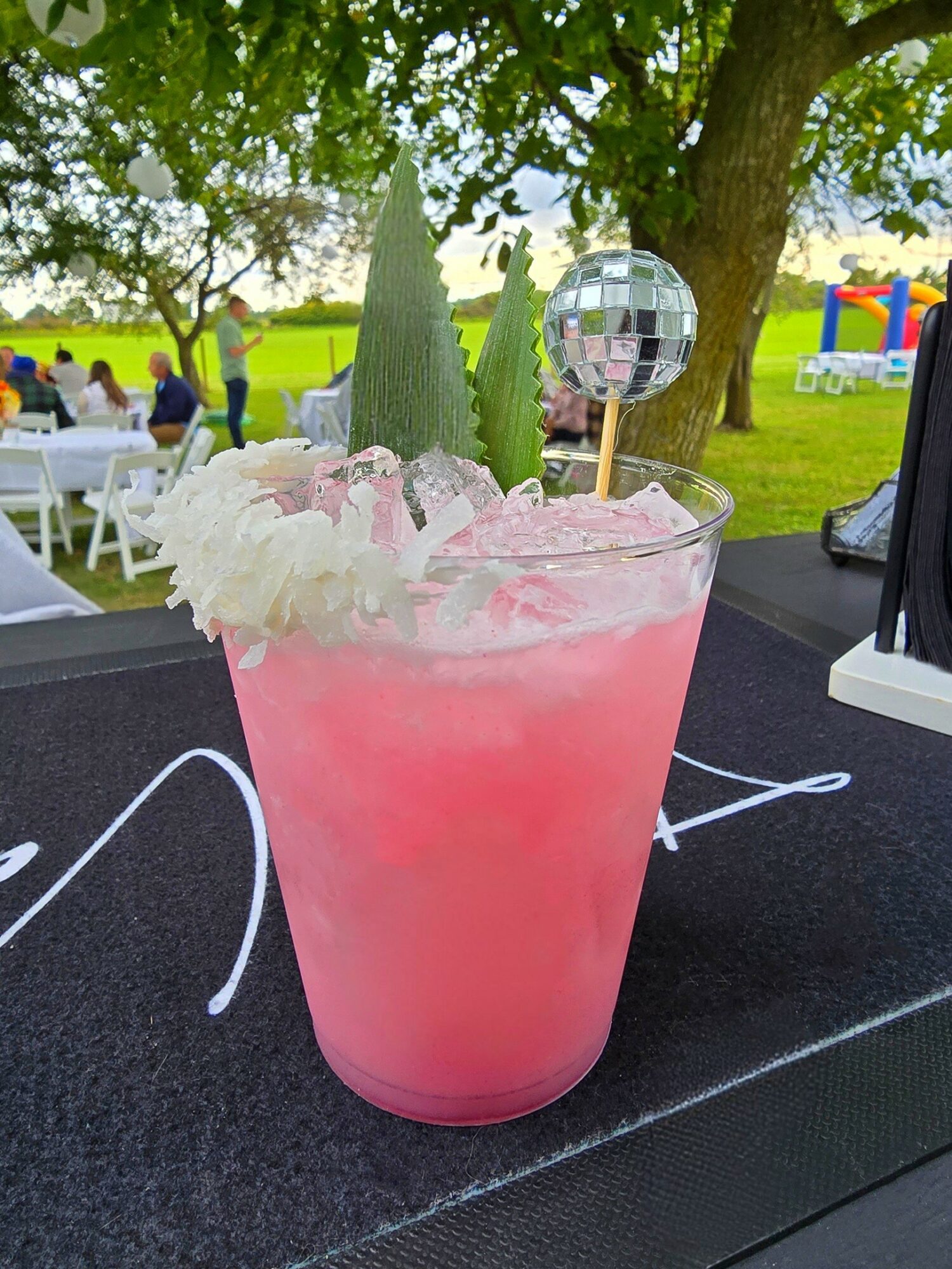 Pink cocktail in a glass with flowers and a mirror ball decoration, outdoors with trees and people in background.