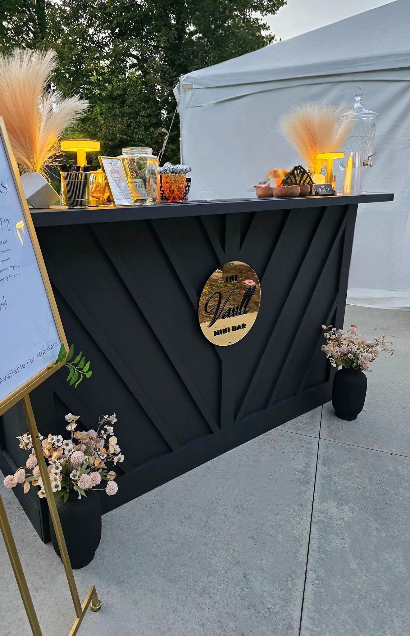 Black bar with a round logo, yellow drink dispensers, and snacks, outdoors near a white tent, with flower pots nearby.