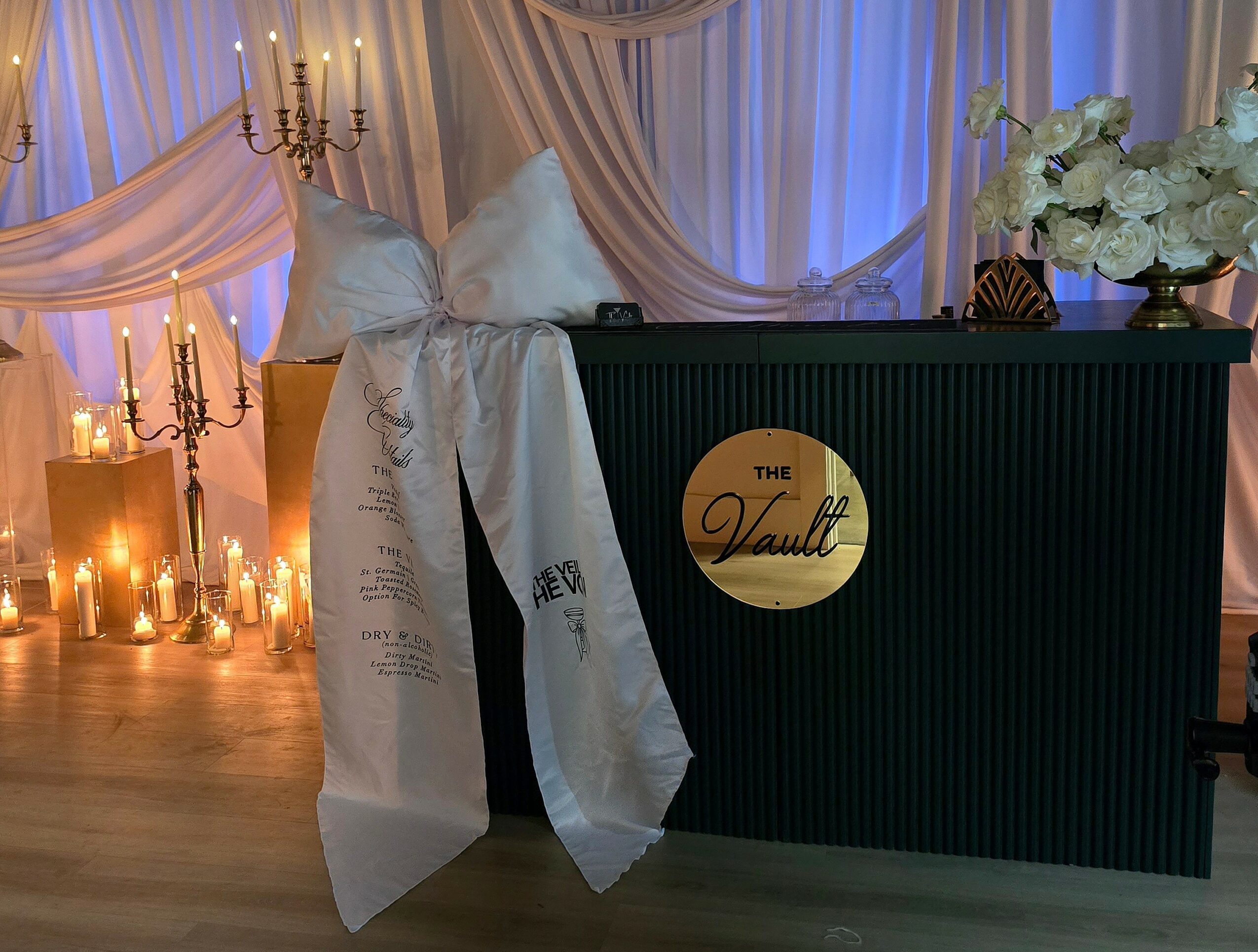 Decorative event setup with candles, draped fabric, and a black counter with a gold logo, flowers, and candles in the background.