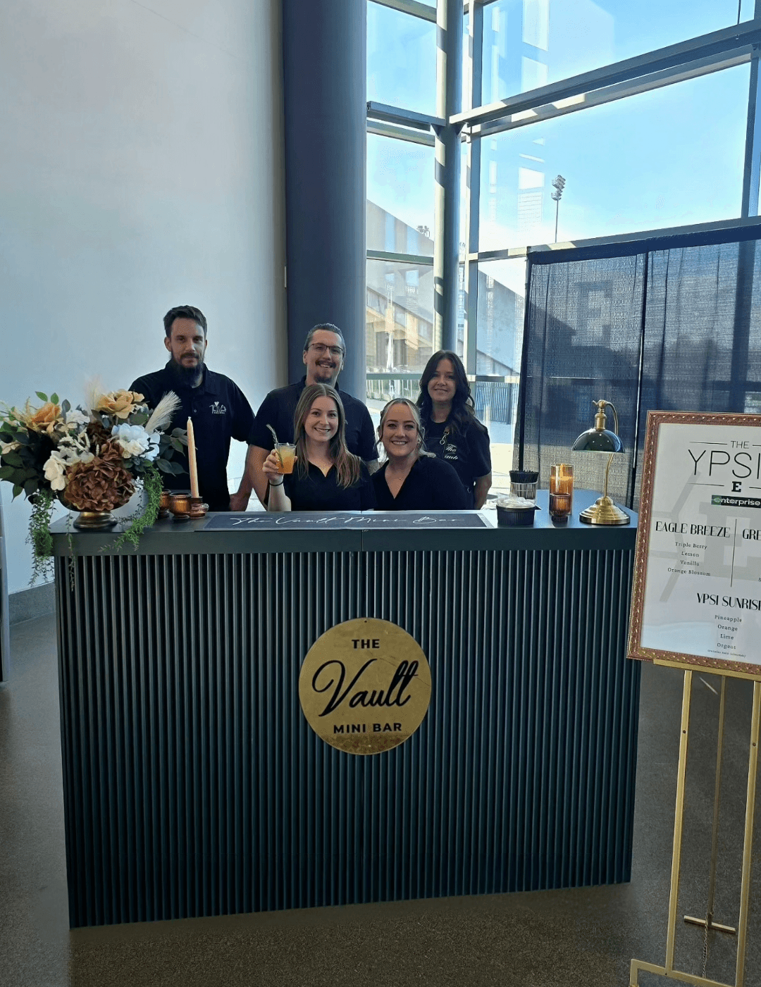 Five people behind a bar counter with a sign reading 'The Vault Mini Bar,' large windows in the background, and a floral arrangement on the left.