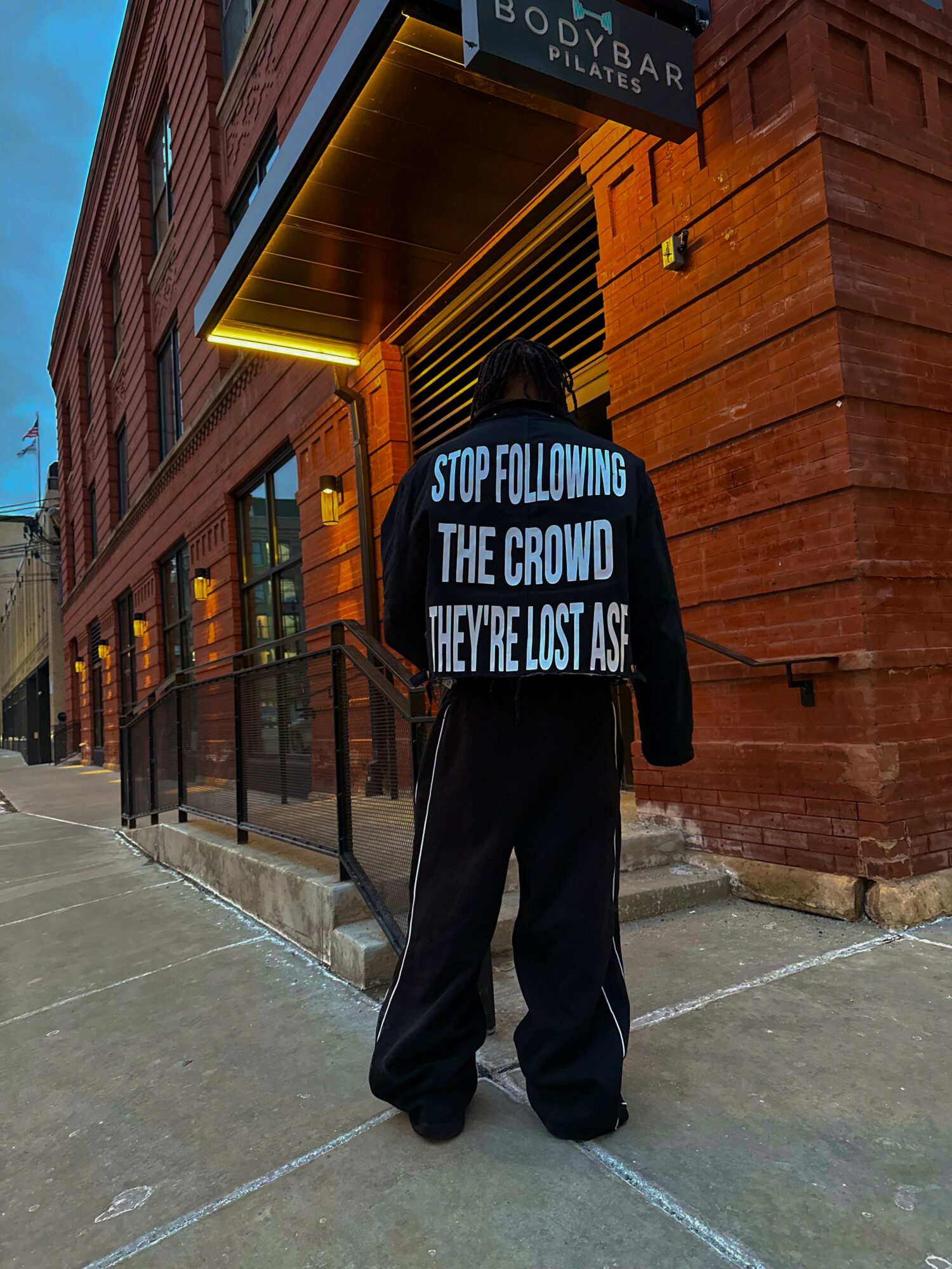 Person standing on sidewalk facing a building with a sign, wearing a jacket with a message on the back.