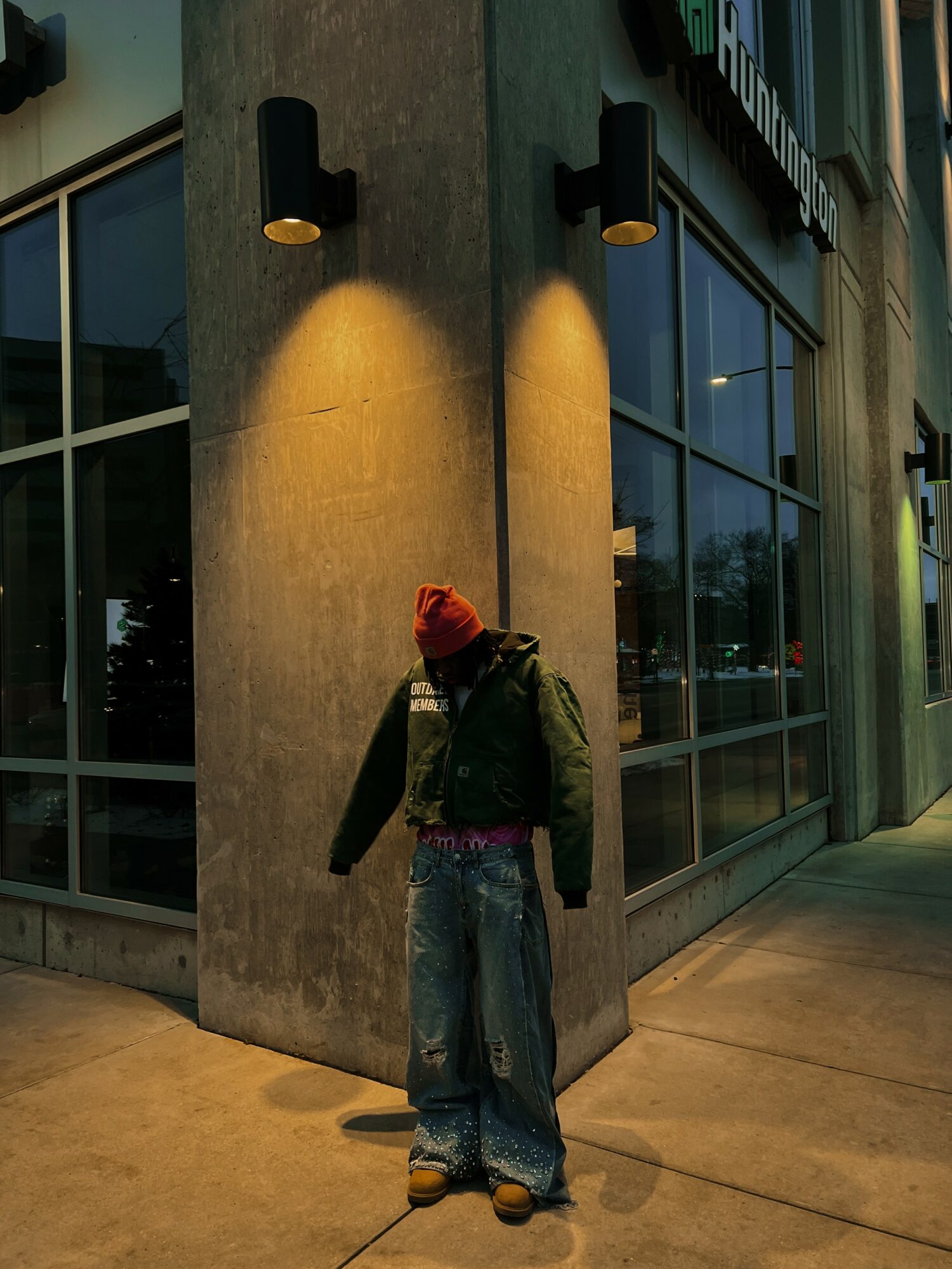 Person standing on sidewalk near building corner, wearing a red hat, green vest, and bag, with large windows behind.
