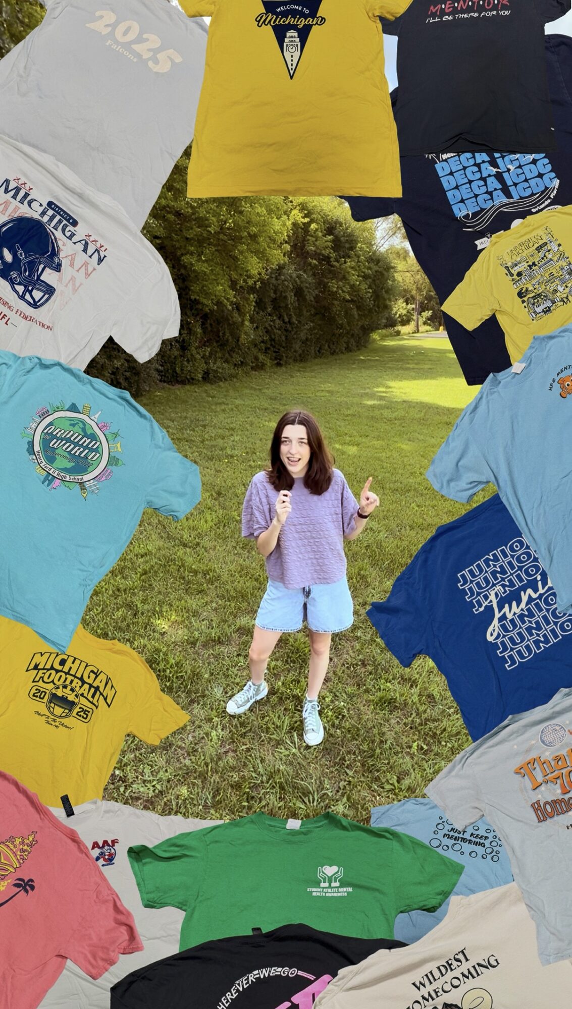 Young girl standing outdoors surrounded by colorful T-shirts laid on grass, holding a drink, smiling, with trees in background.