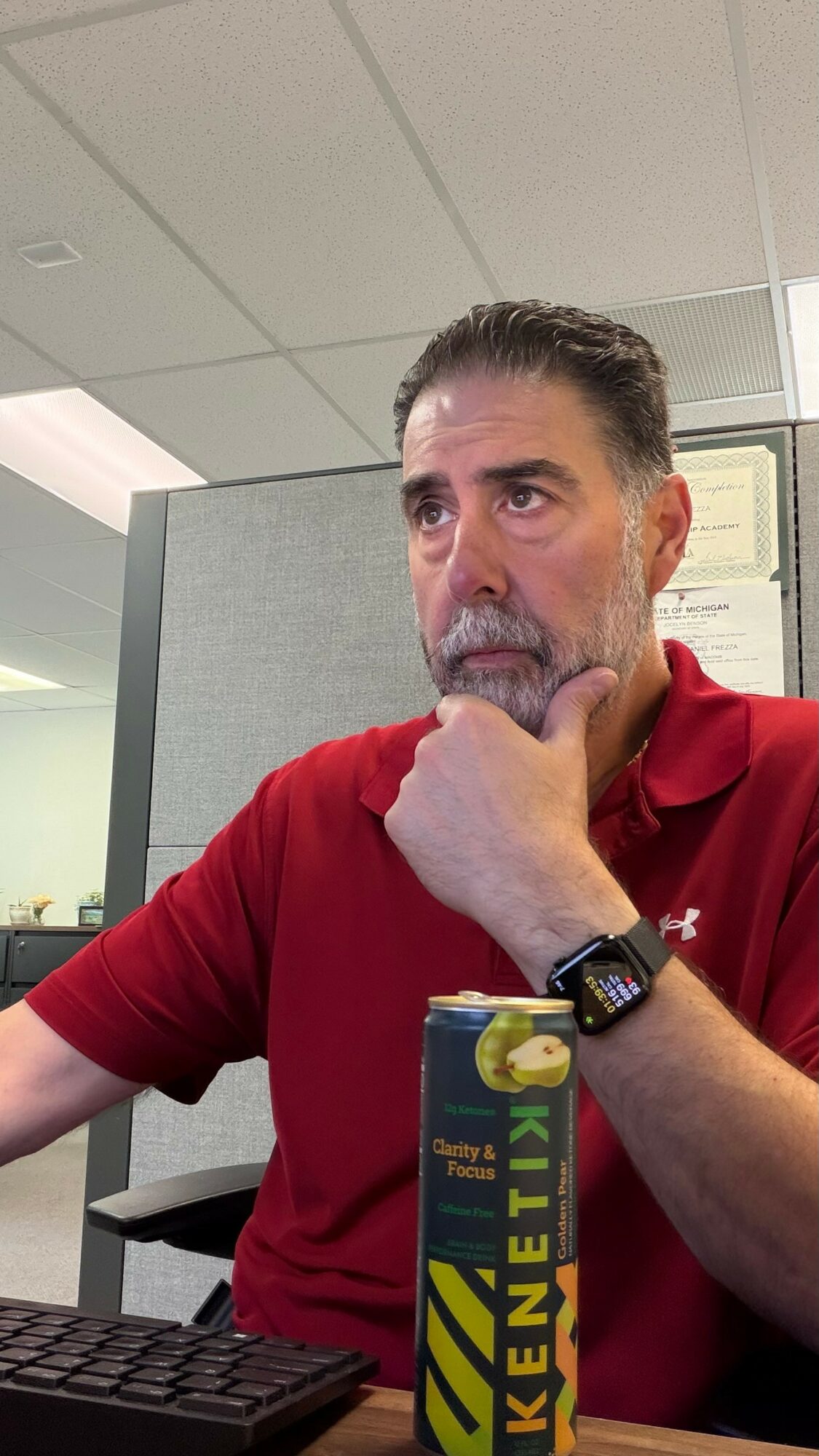 Man with gray hair and beard wearing a red polo shirt, sitting at a desk in an office, resting his chin on his hand.