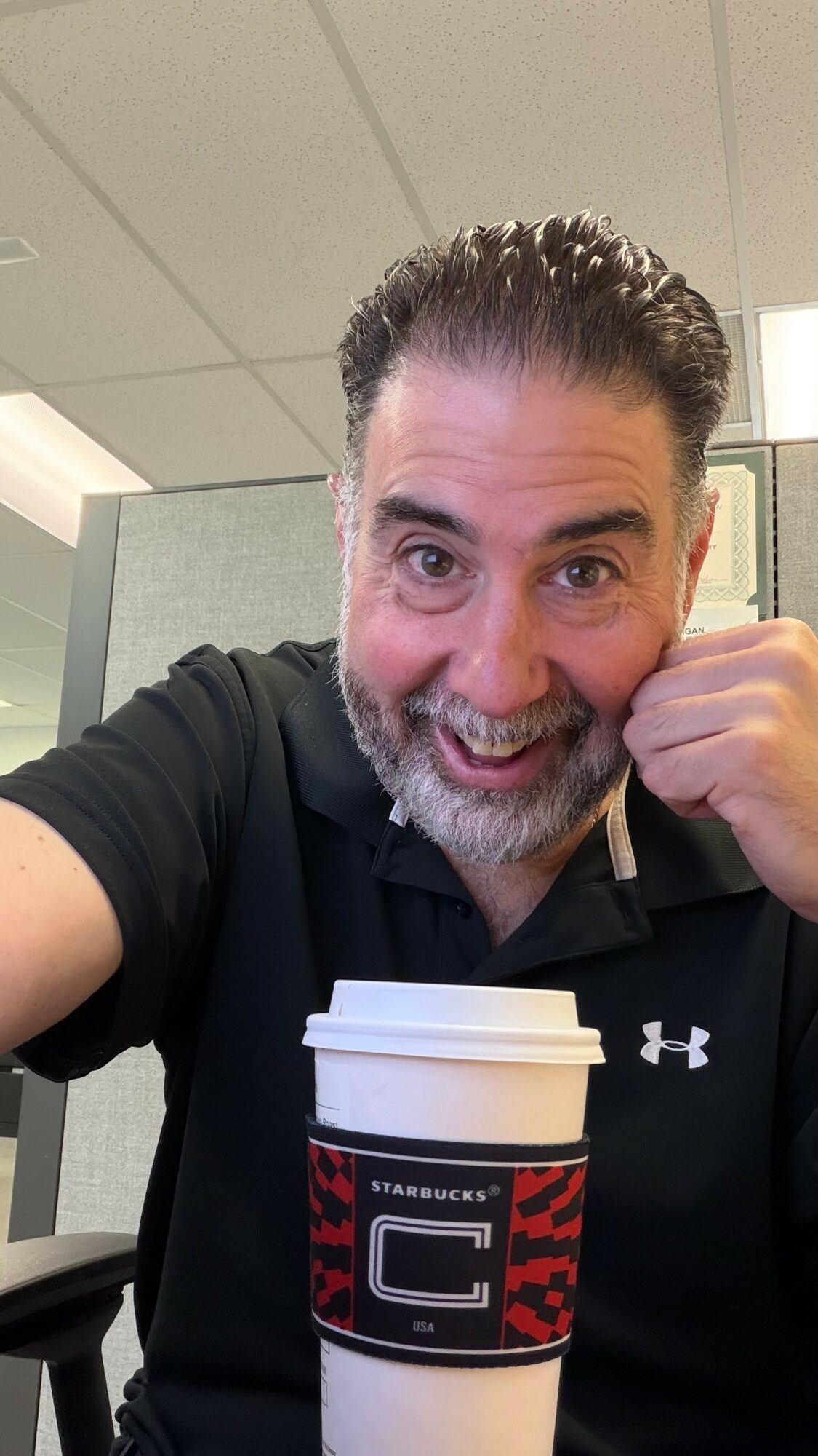 Man smiling with a coffee cup in an indoor setting, wearing a black polo shirt, raising his fist.