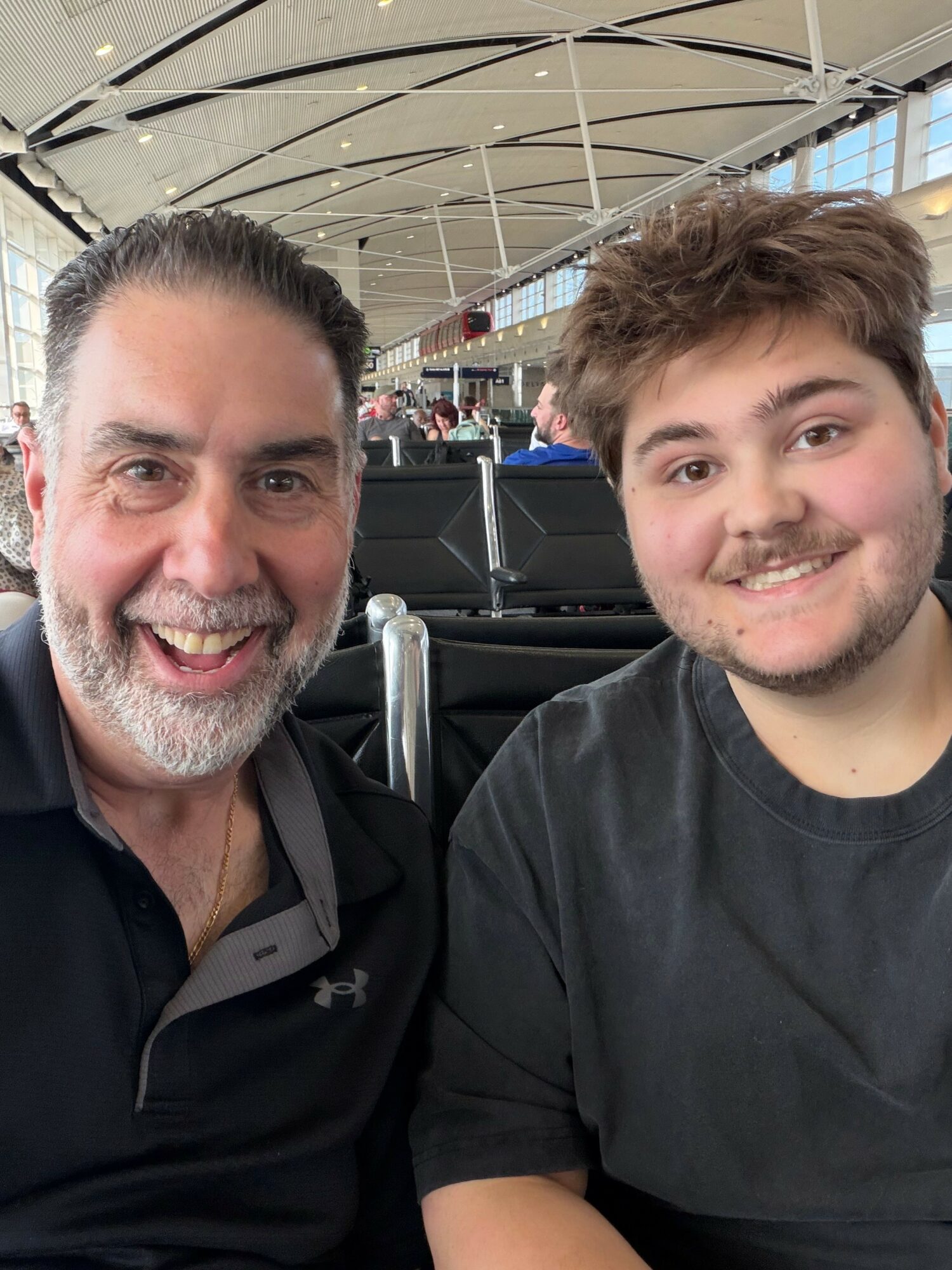 Two smiling males sitting in an airport terminal, with a curved ceiling and large windows in the background.