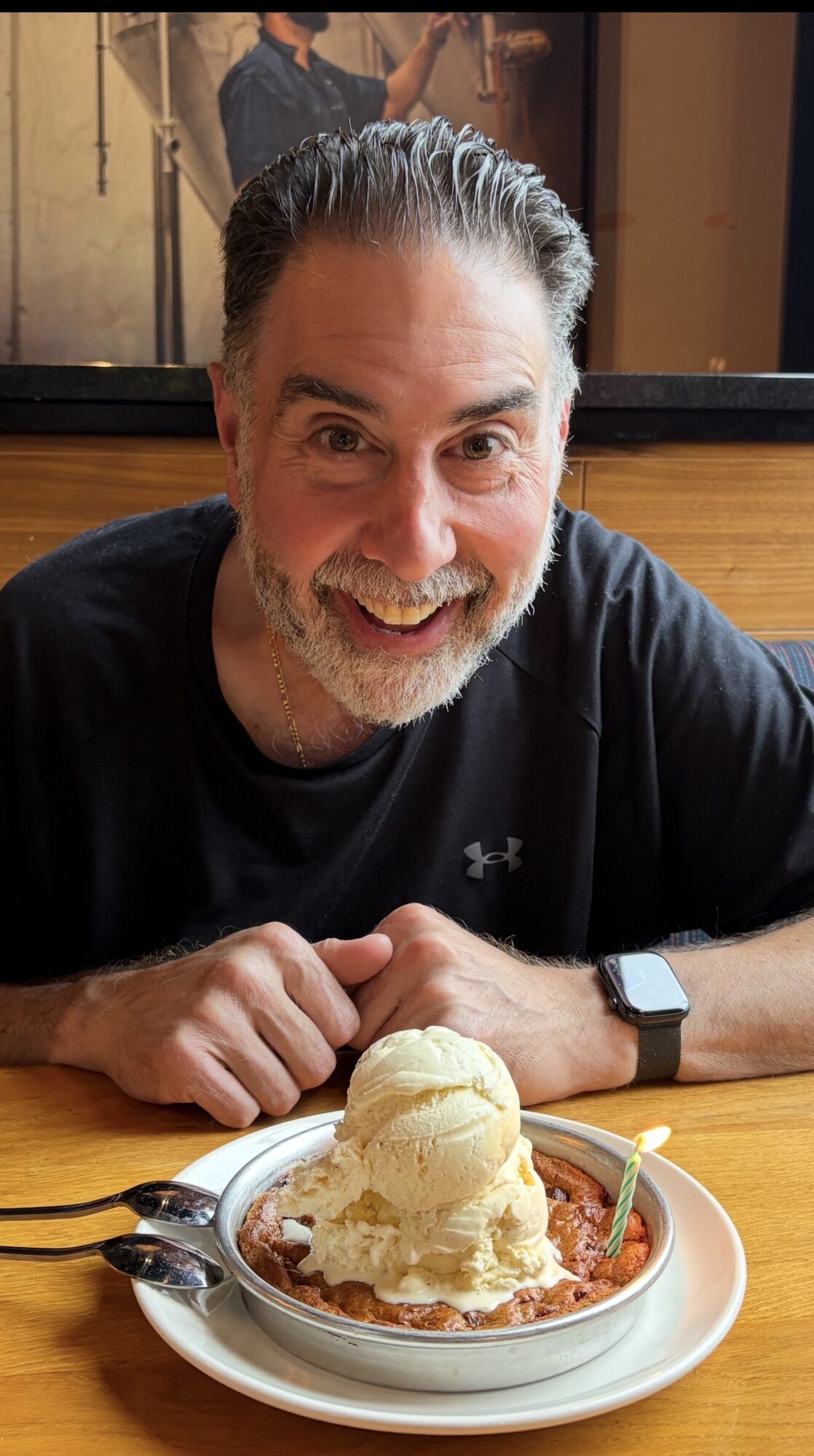 Man with gray hair and beard smiling at a dessert with ice cream and sauce, sitting at a table.