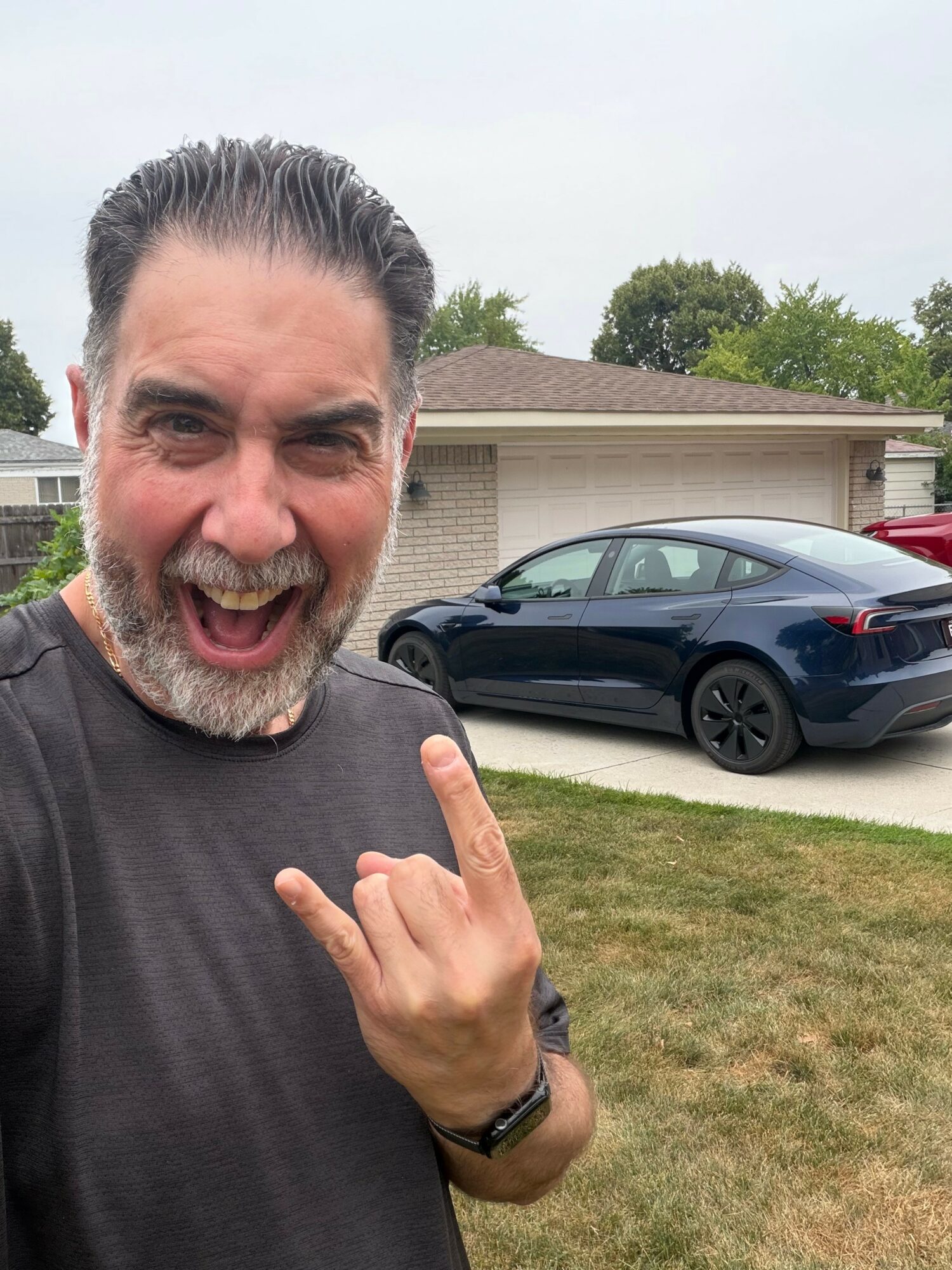 Man smiling and making a rock hand gesture outdoors with a car and house in the background.
