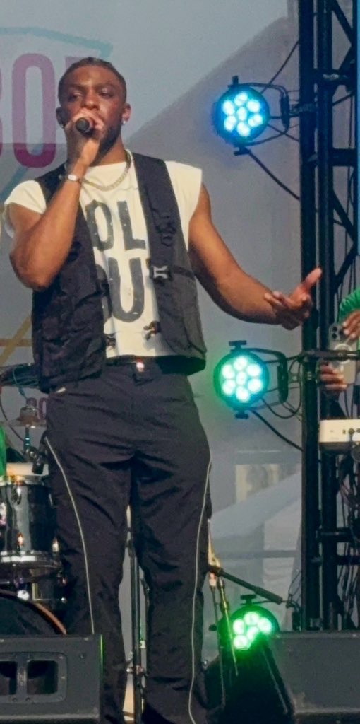 Male singer holding microphone on stage with blue and green stage lights, wearing a vest and pants.