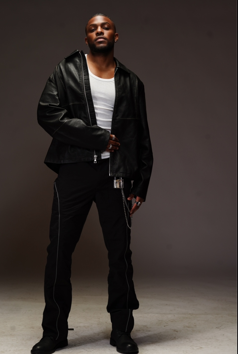Man standing in black jacket, white shirt, and black pants against a dark background.