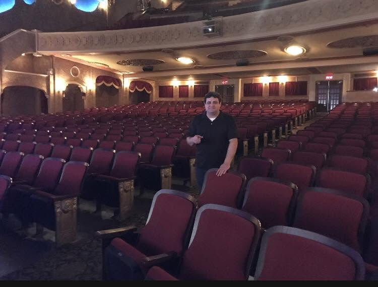 Person standing in an empty theater with rows of red seats and ornate ceiling details.