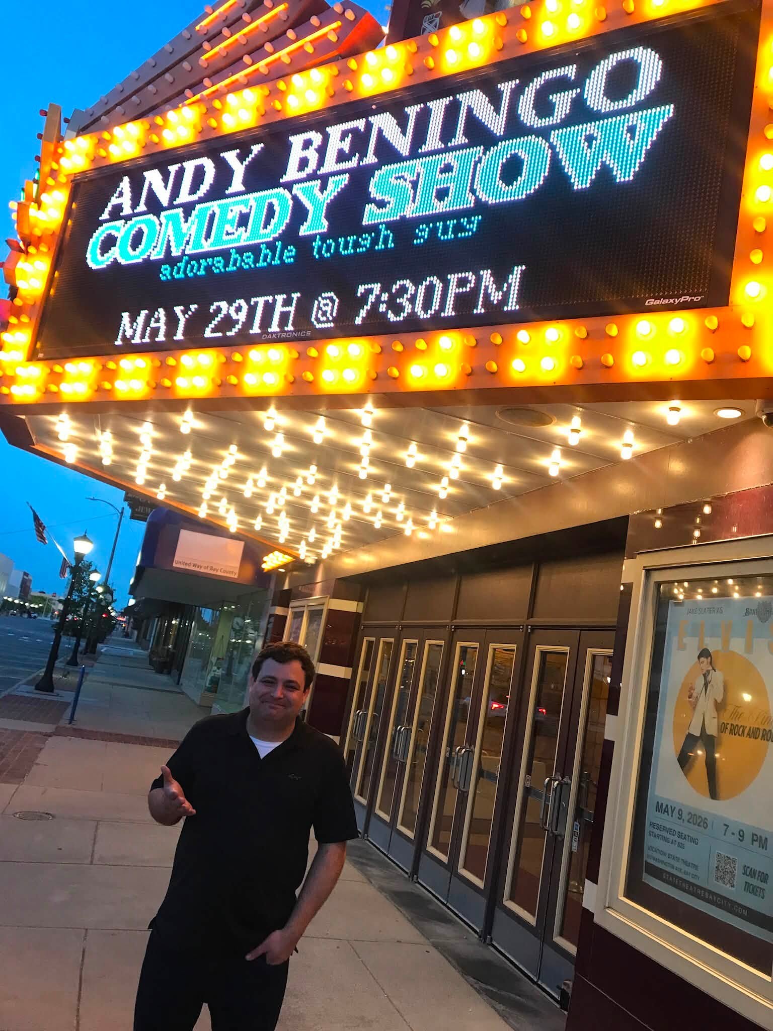 Person standing outside a theater with a marquee advertising Andy Beningo comedy show on May 29th at 7:30 PM, illuminated lights, evening sky.