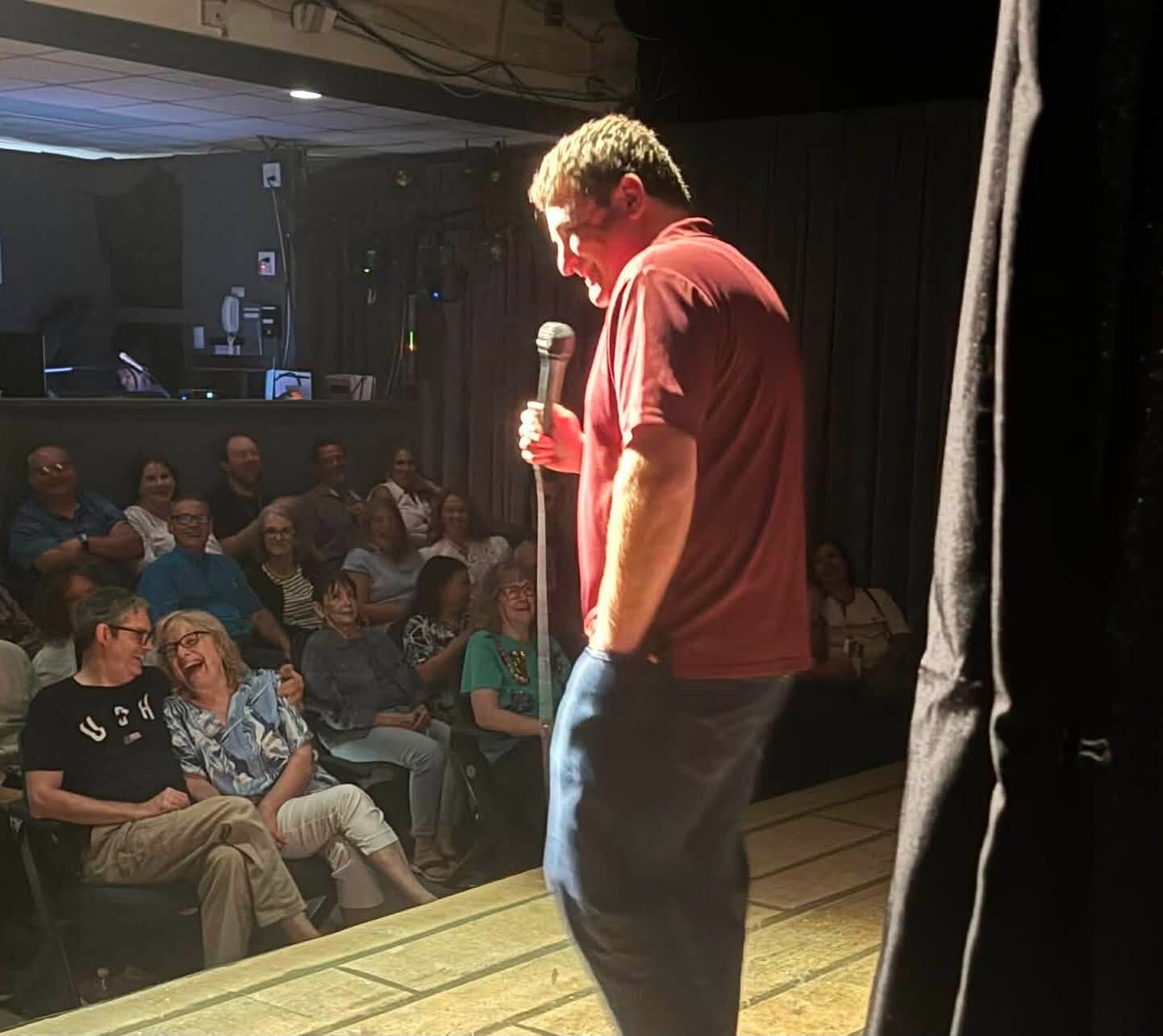 A man stands on stage holding a microphone, facing an audience in a dark room with black curtains.