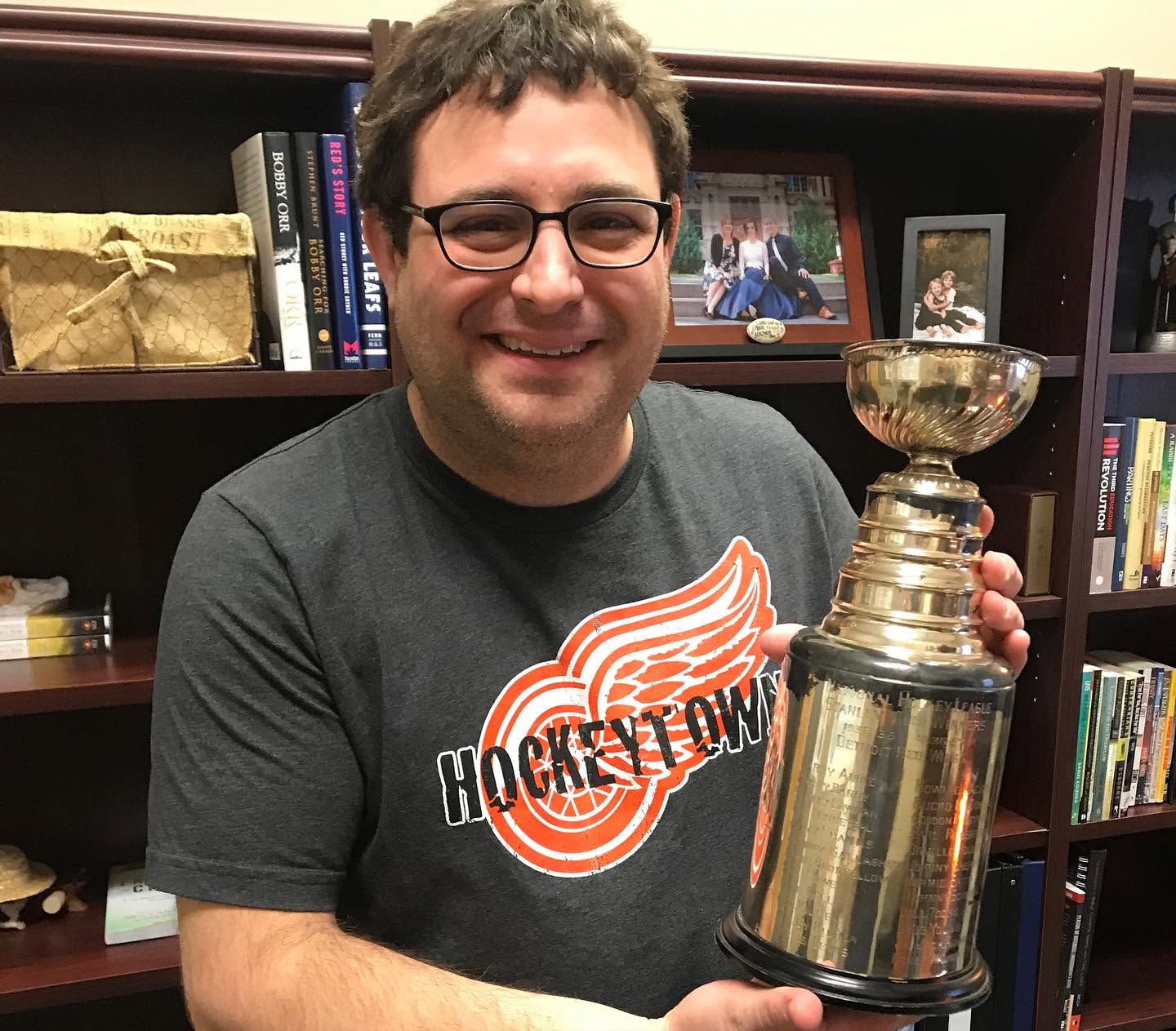Man with glasses smiling, holding a large trophy, standing in front of a bookshelf with books and framed photos.