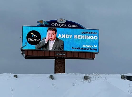 Billboard advertising comedian Andy Beningo at Odawa Casino, with snow-covered ground below, blue sky background.