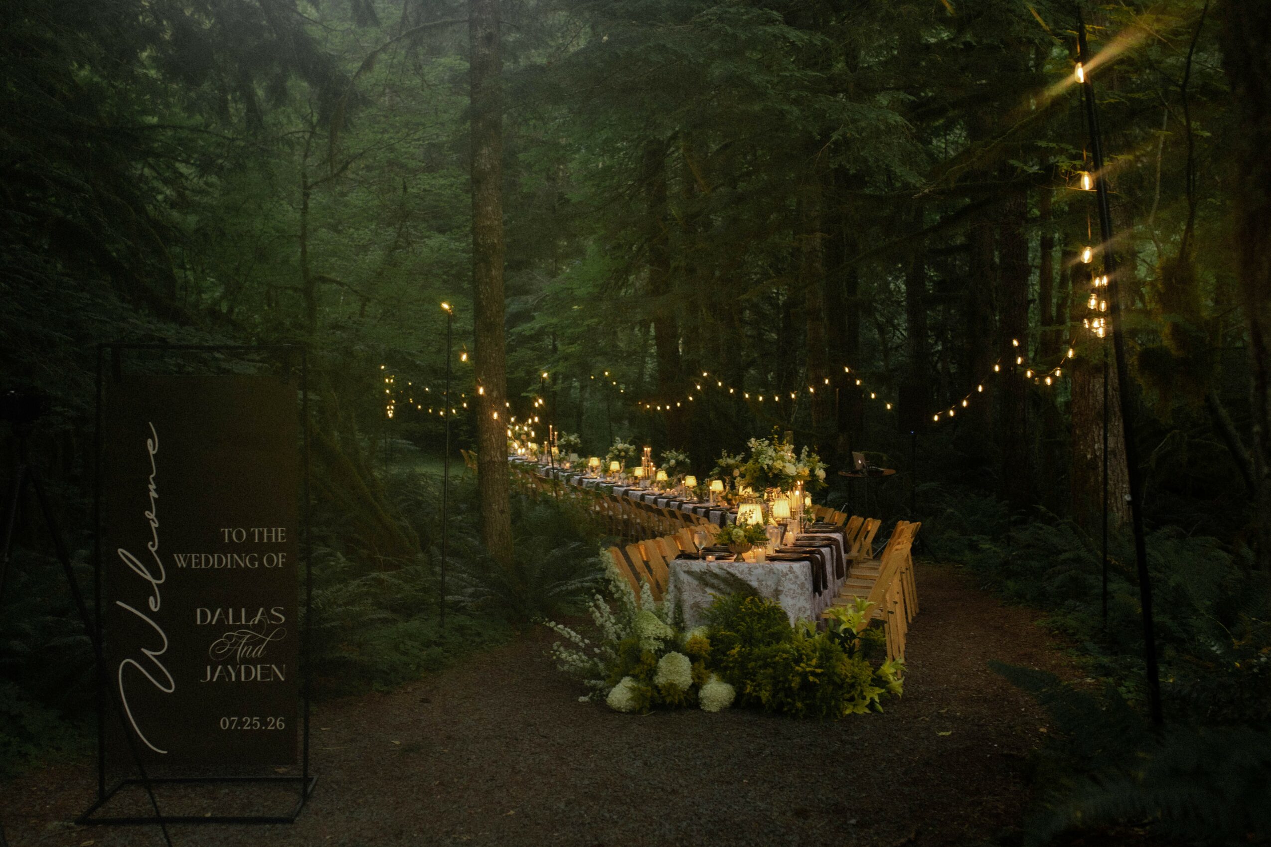 Outdoor wedding reception setup in a forest with string lights, a decorated table, and floral arrangements, nighttime scene.