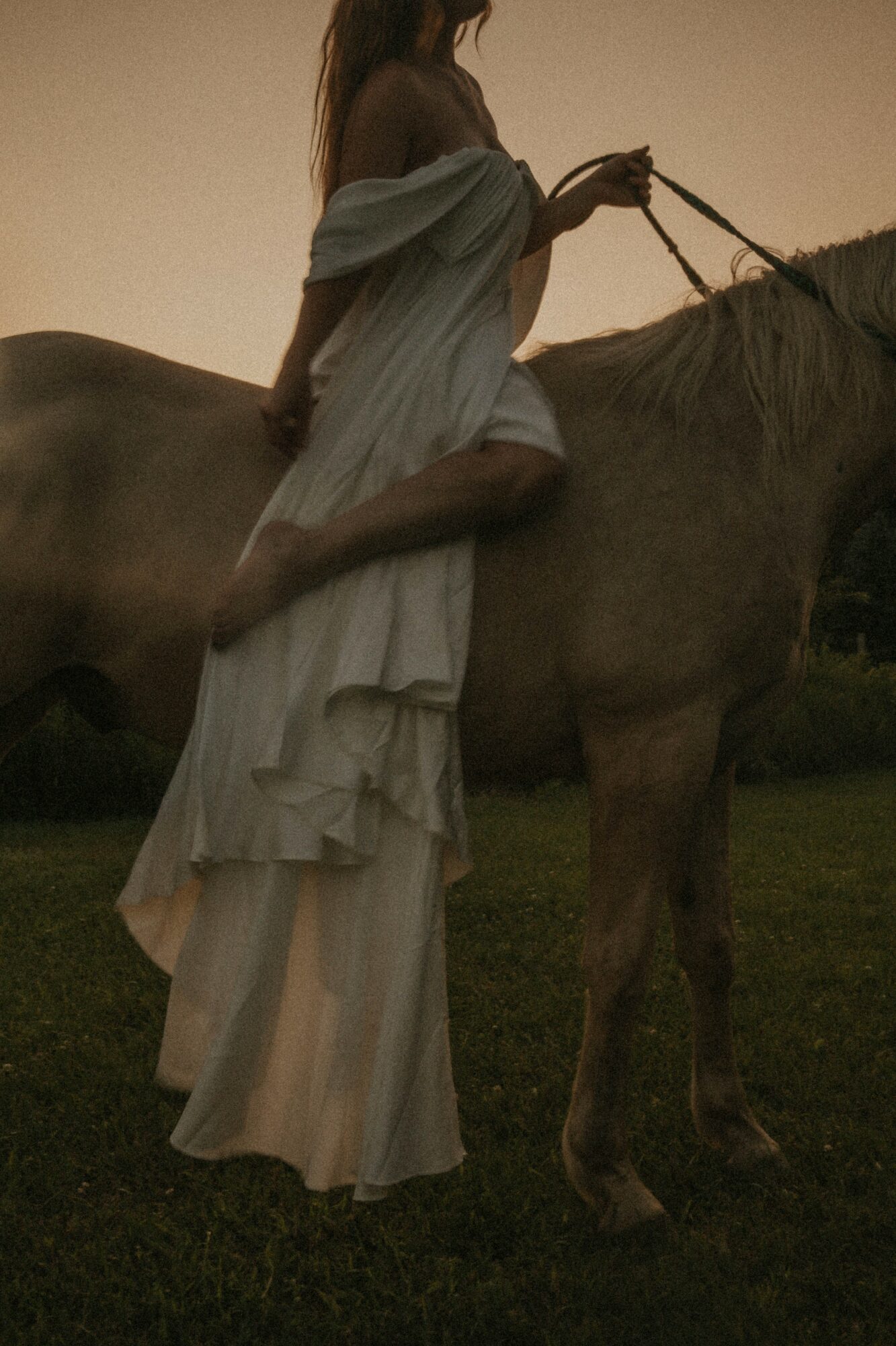 Person in flowing dress sitting on a horse outdoors during sunset, holding the reins.