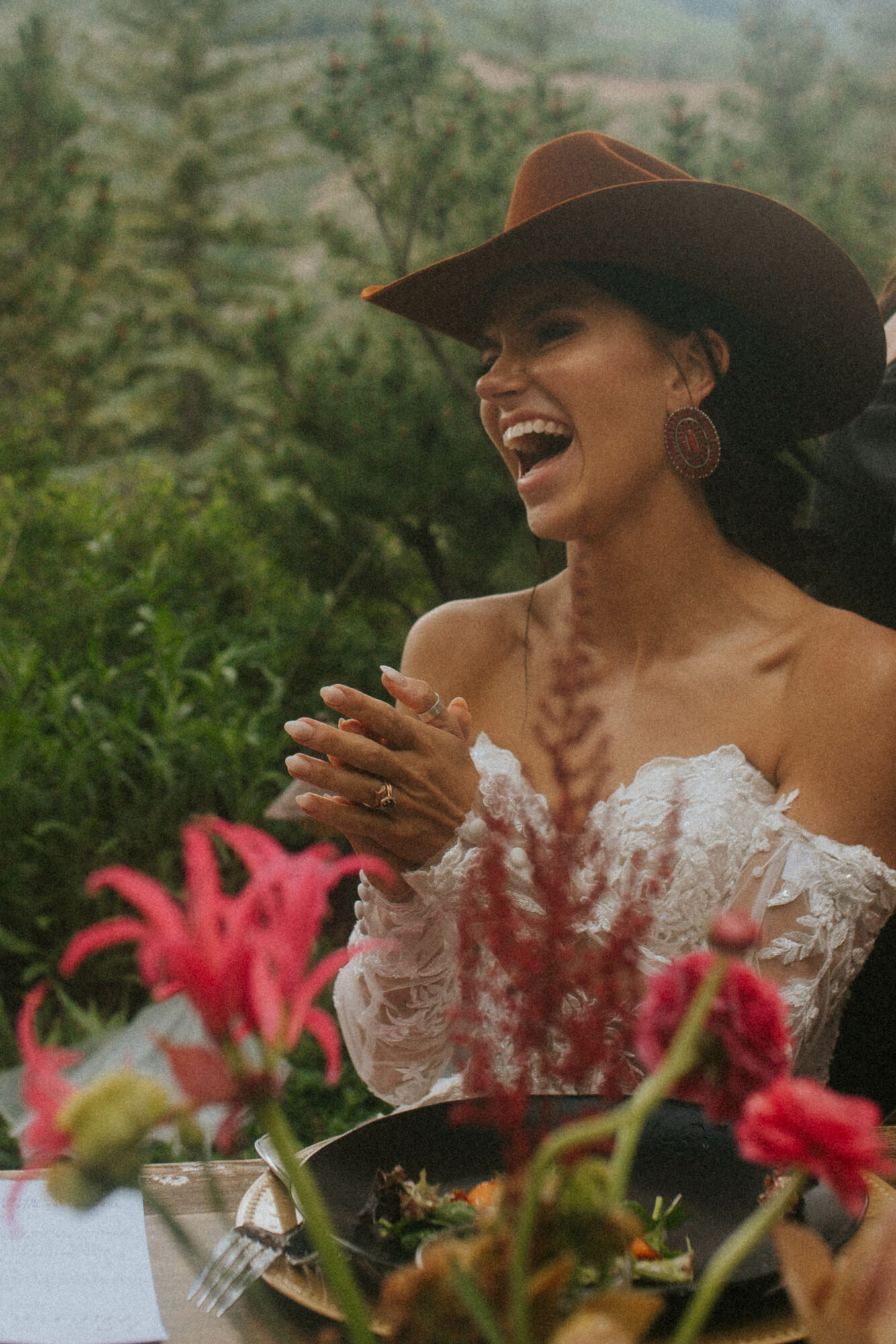 Woman wearing a wide-brimmed hat and lace dress, smiling outdoors with greenery and pink flowers in foreground.