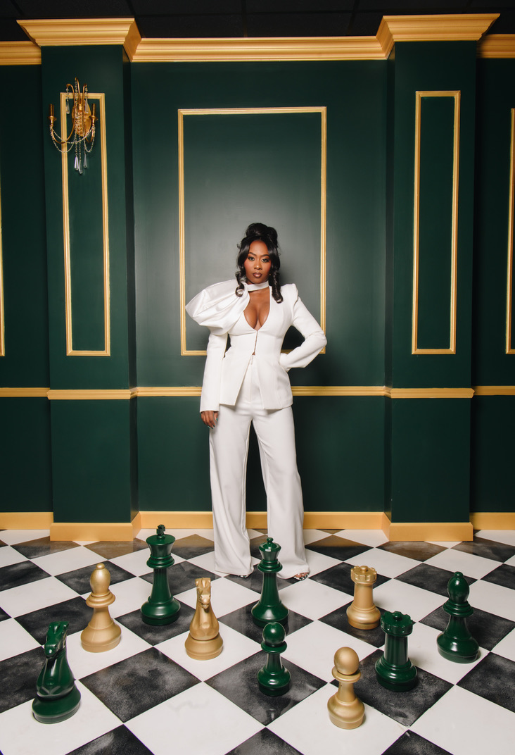 Woman in white suit standing on a chessboard-patterned floor against a green wall with gold trim, posing with one hand on her hip.
