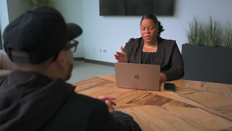 Woman speaking during a video call, sitting at a table with a laptop, in a modern office setting.