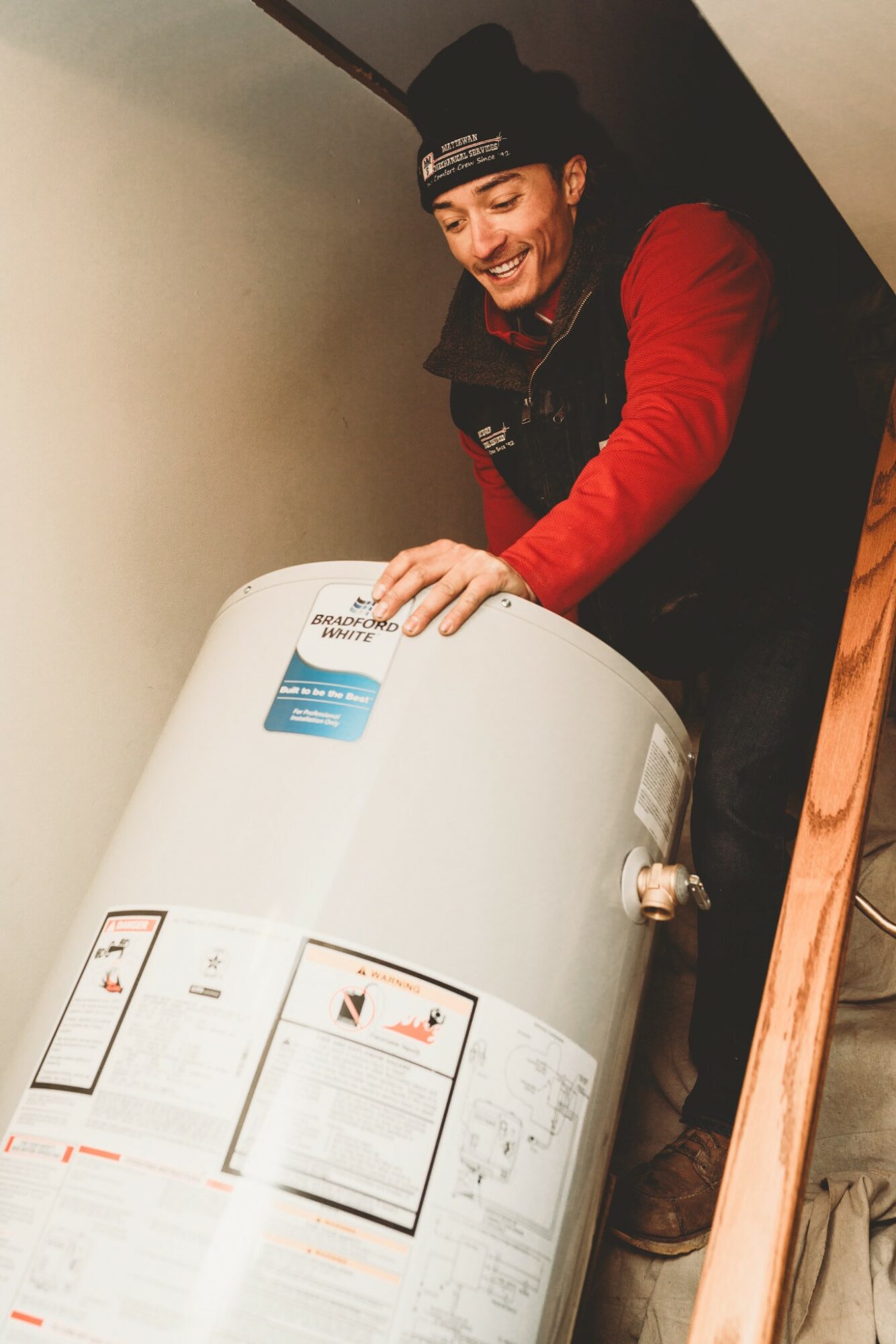 Person smiling, wearing a black beanie and red jacket, leaning over a white water heater indoors.