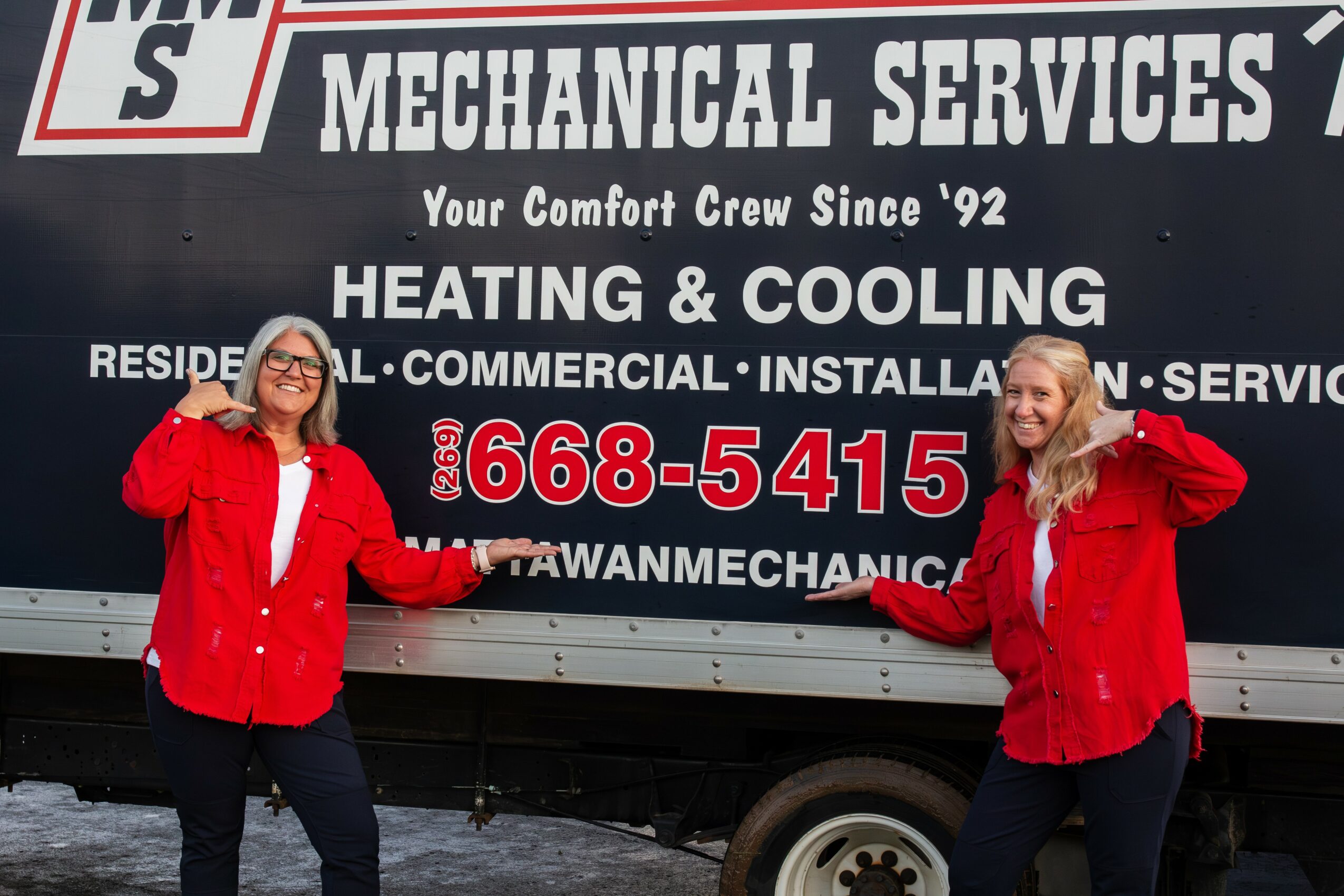 Two women in red jackets standing in front of a truck with a sign for mechanical services, holding a long object between them.