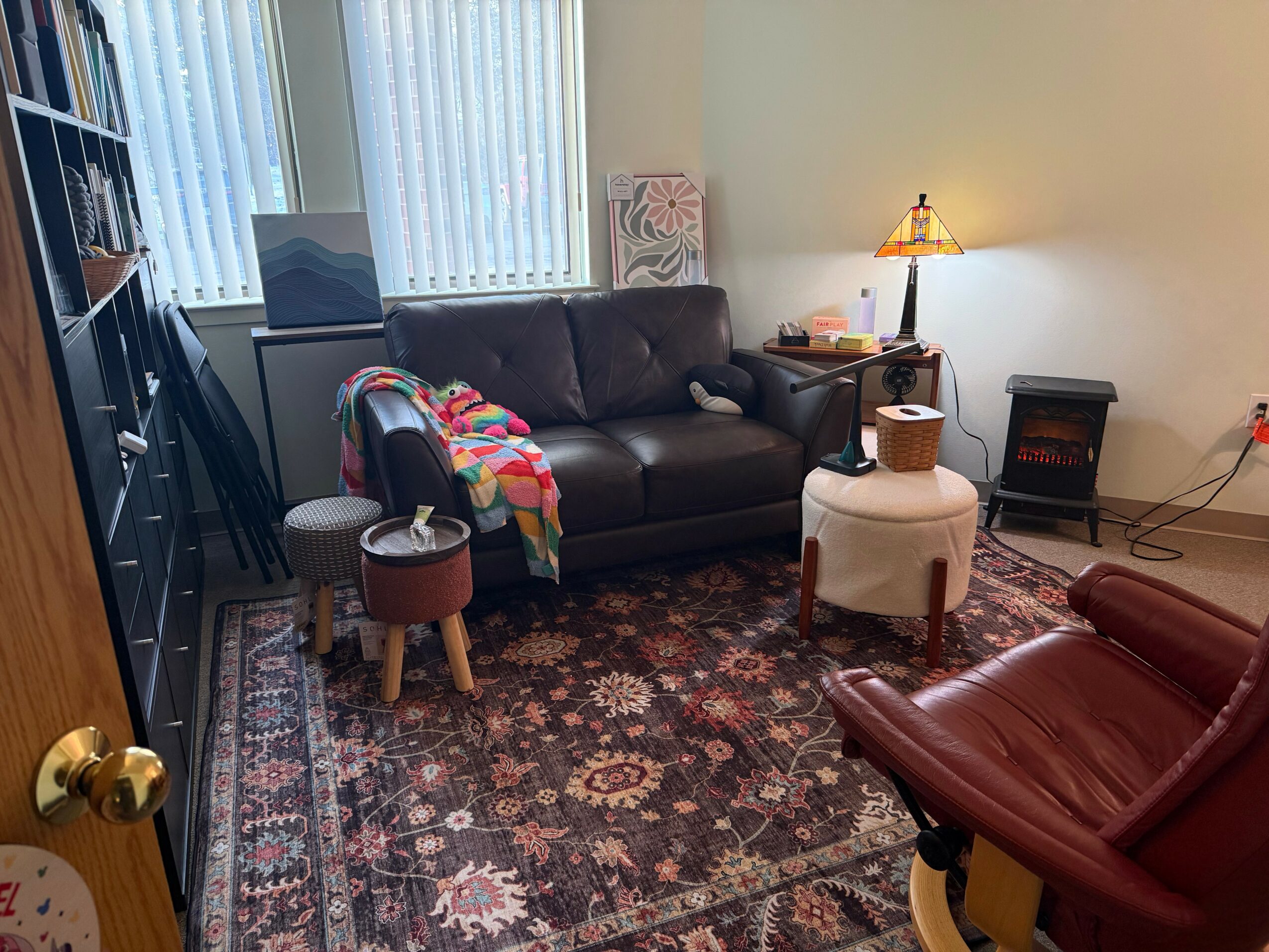 Living room with a black sofa, a red armchair, a small white ottoman, and a patterned rug. Window with blinds, lamp, and bookshelf.