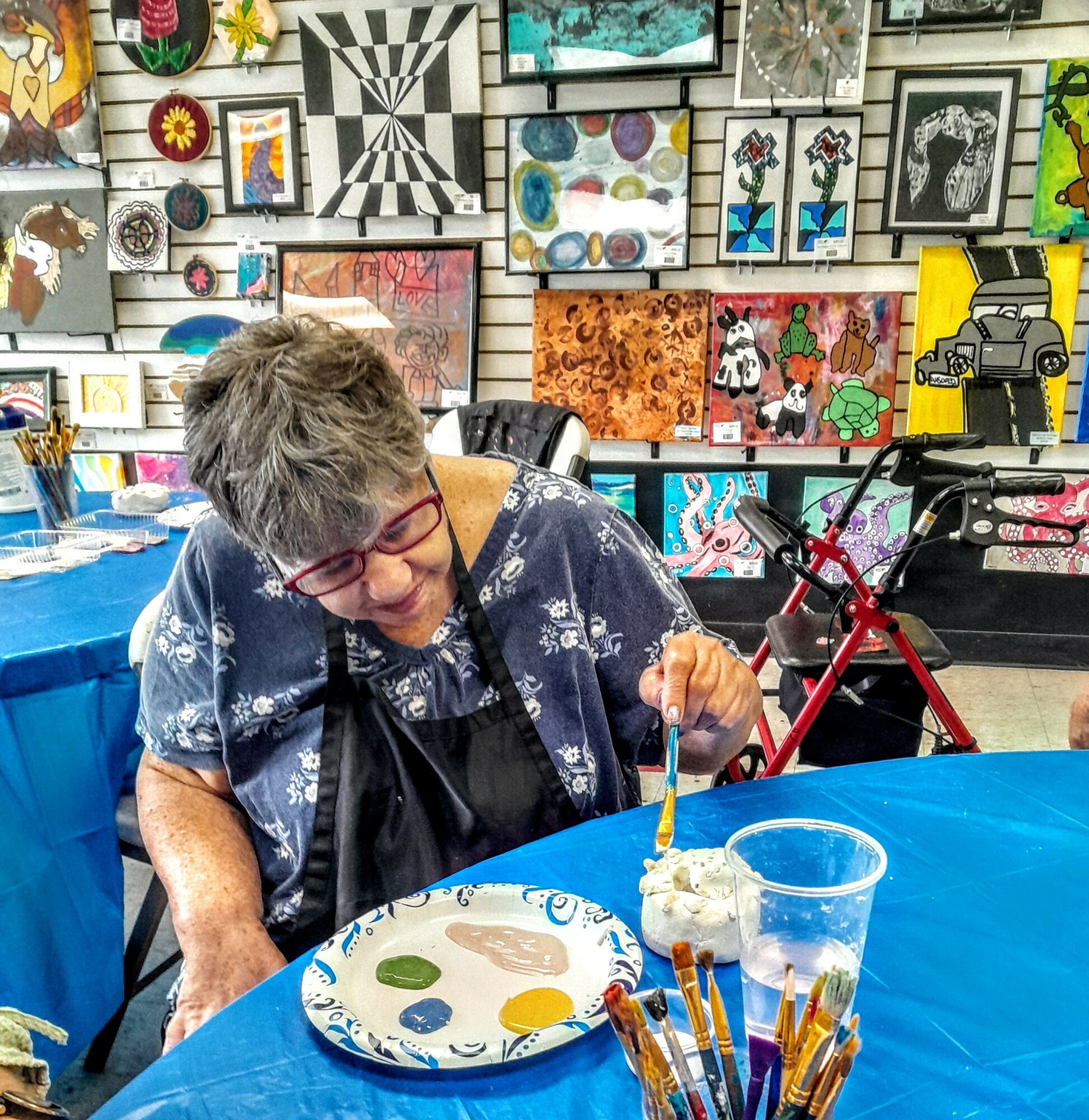 Older woman with short gray hair and glasses, sitting at a table with art in the background, holding a skewer with food.