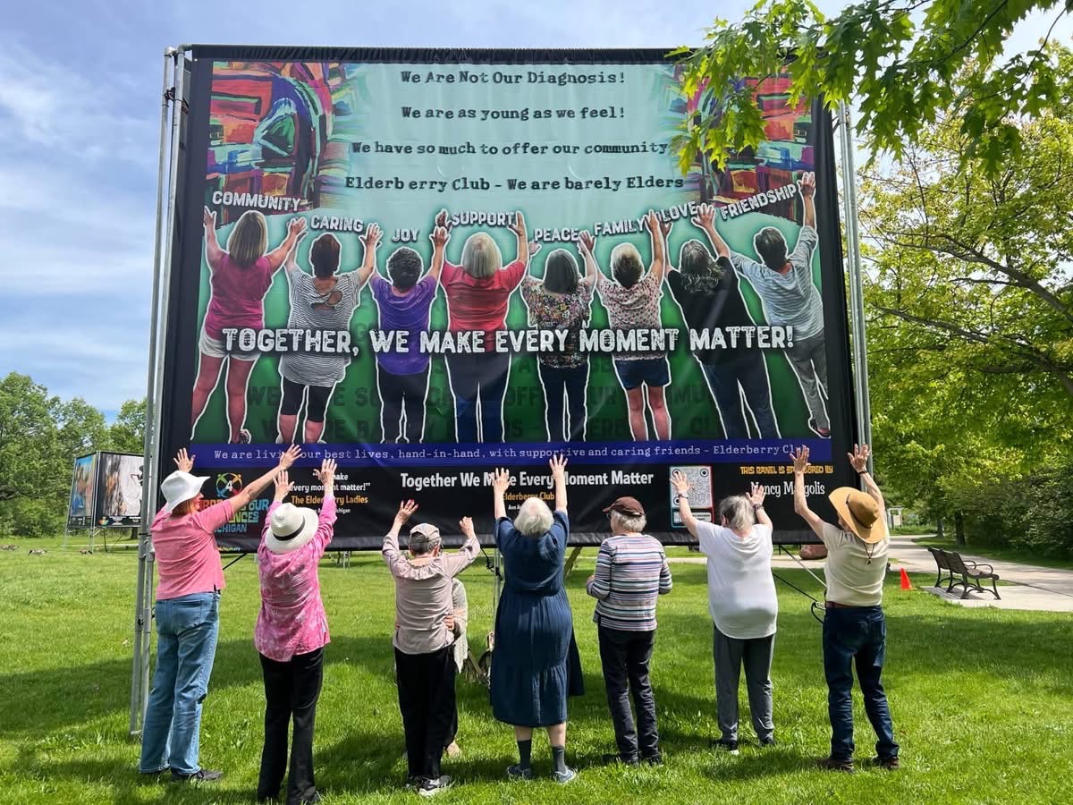 Group of people standing outdoors, facing a large screen displaying colorful artwork and text, with trees and a park in the background.