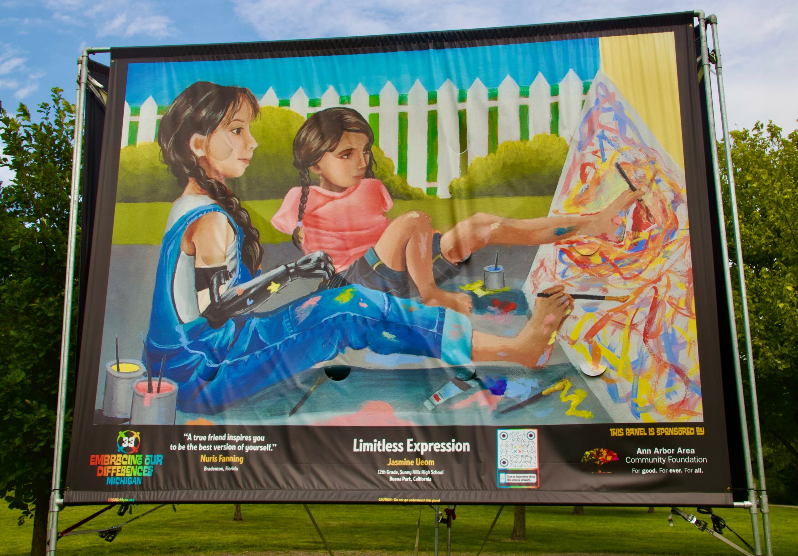 Two children sit on a blanket outdoors with a white picket fence and trees in the background.