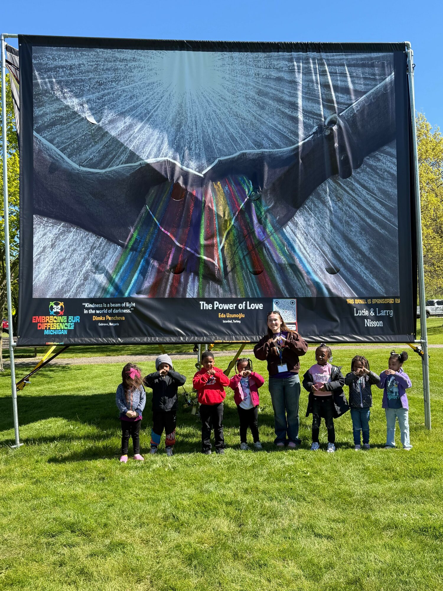 Group of children and adults standing on grass in front of a large outdoor screen displaying a mountain scene with rainbow light effects.