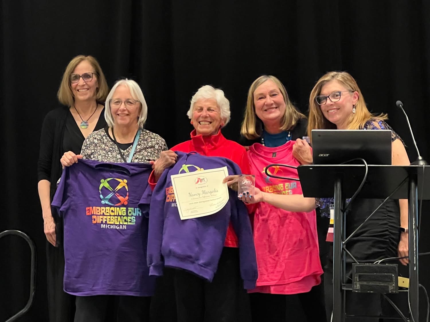 Five women standing together, smiling, with one holding a purple shirt and a certificate, in front of a black curtain.