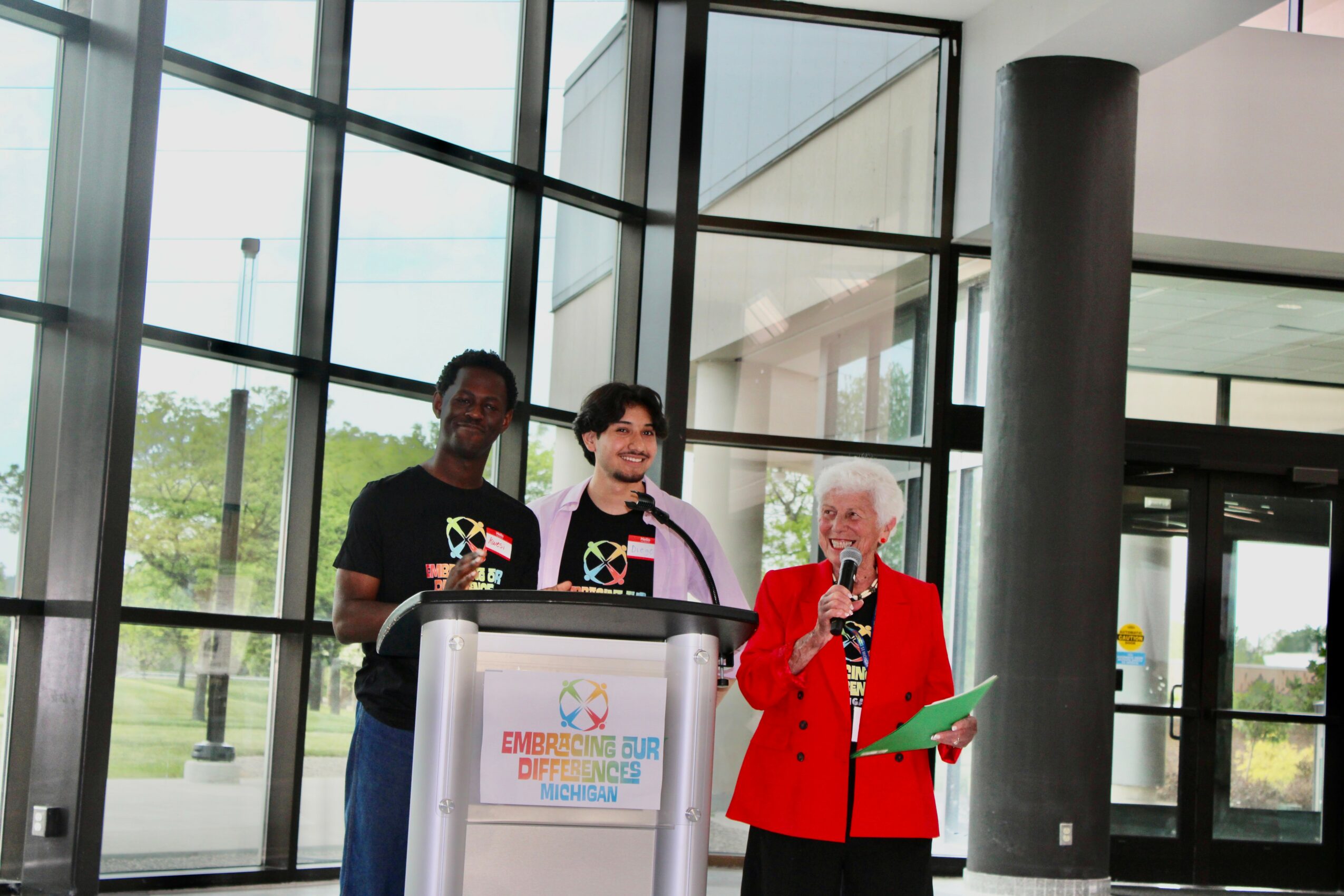 Three people standing behind a podium in a modern building with large glass windows, one speaking into a microphone.