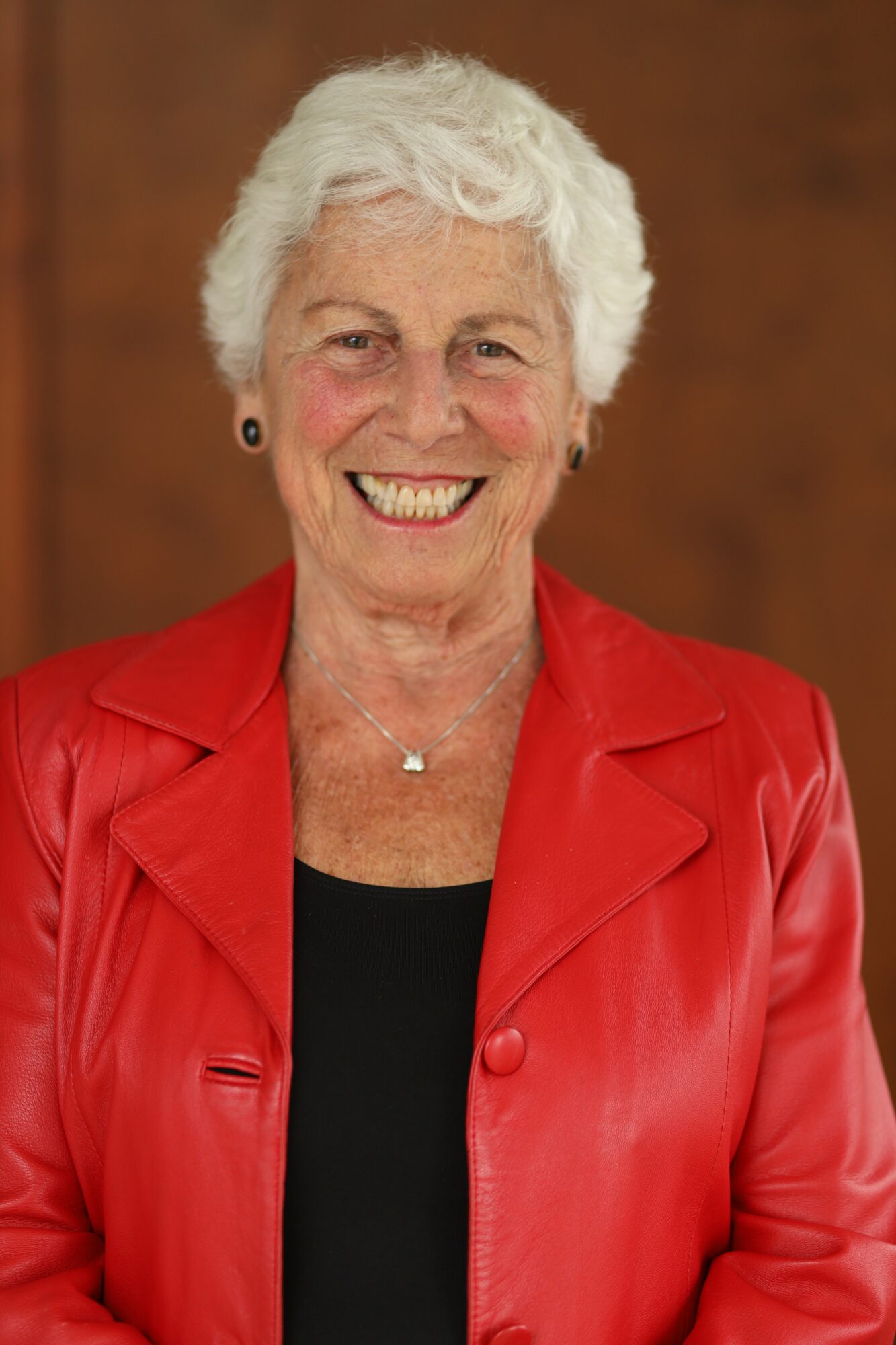 Smiling elderly woman with short white hair, wearing a red jacket and black top, against a brown background.