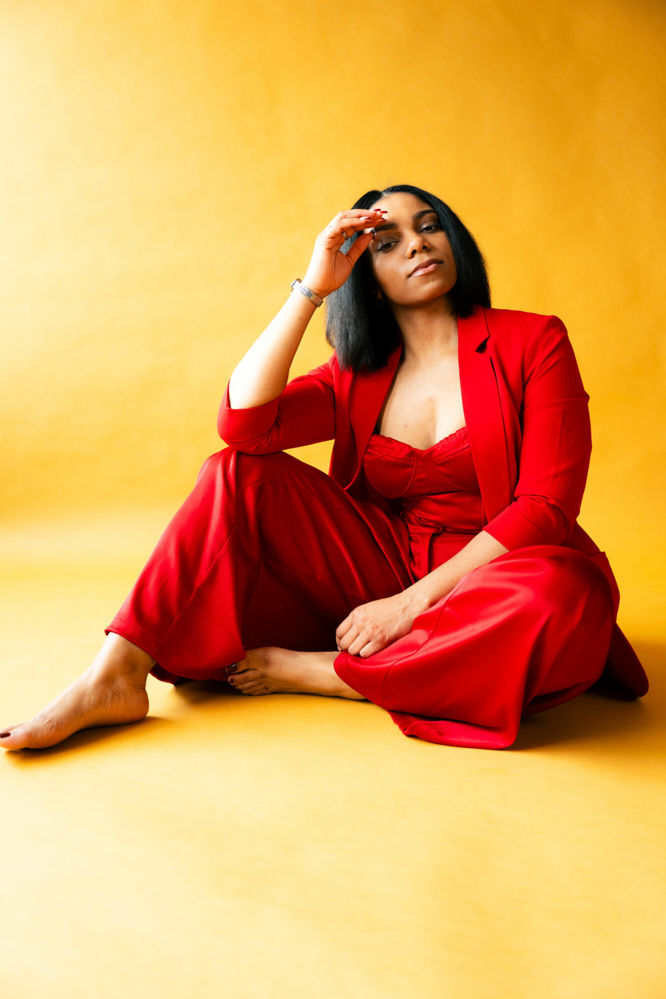 Woman in red jumpsuit sitting on yellow background, looking at camera, with one hand near her face.
