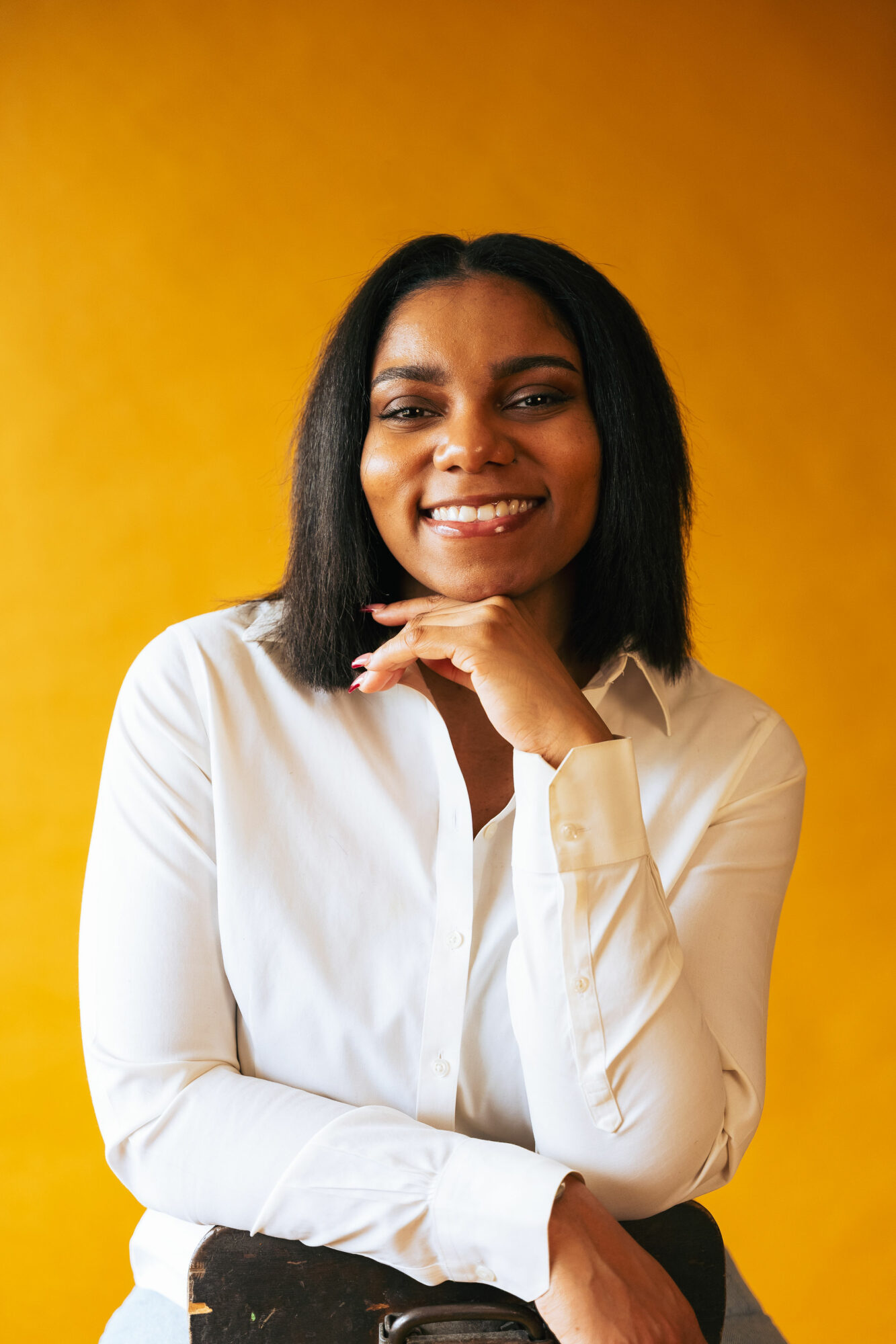 Smiling woman with shoulder-length black hair wearing a white shirt against a yellow background.