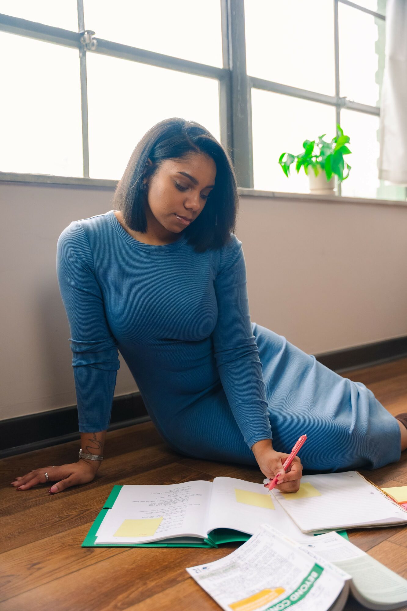 Woman in blue dress sitting on floor, writing in open book, near window with potted plant, natural light.