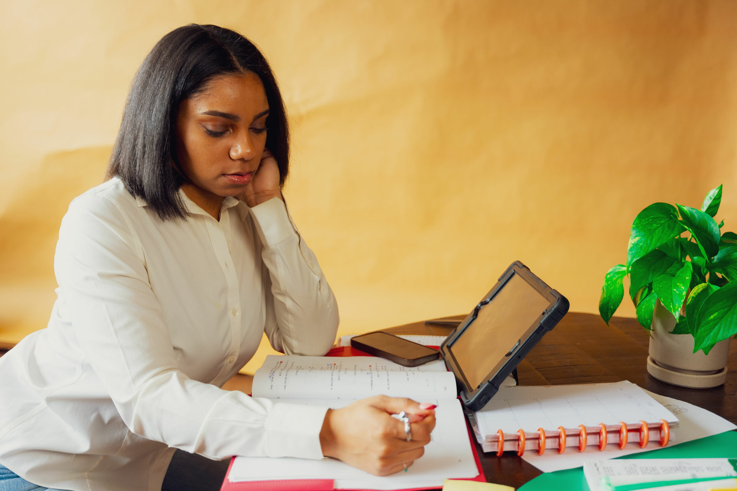 Woman with shoulder-length black hair reading a book at a desk with a tablet, notebook, and plant nearby.