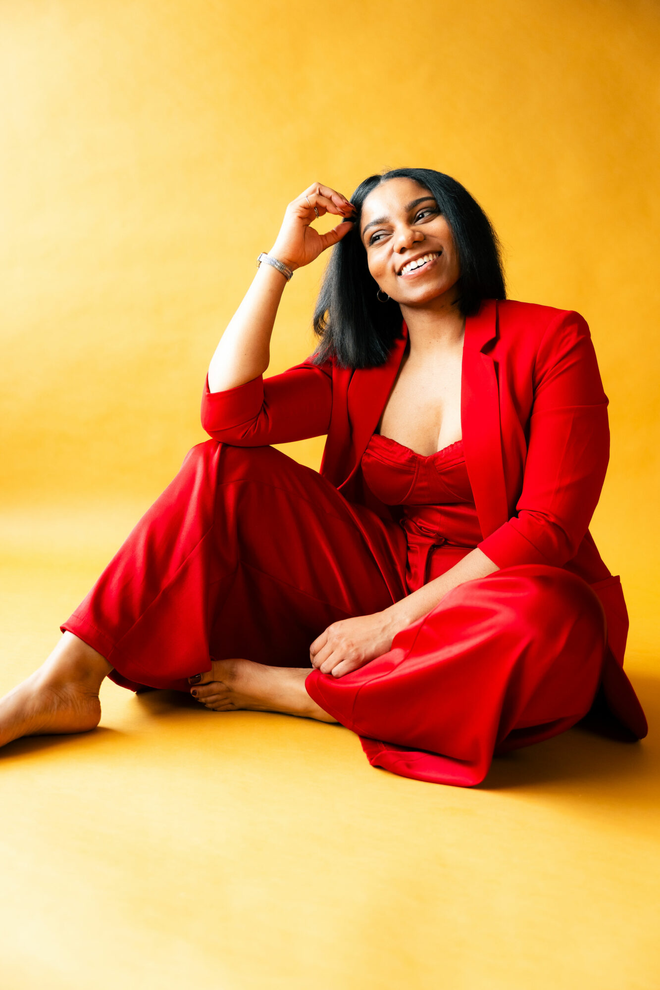 Woman sitting on the floor in a red outfit, smiling, with a yellow background.