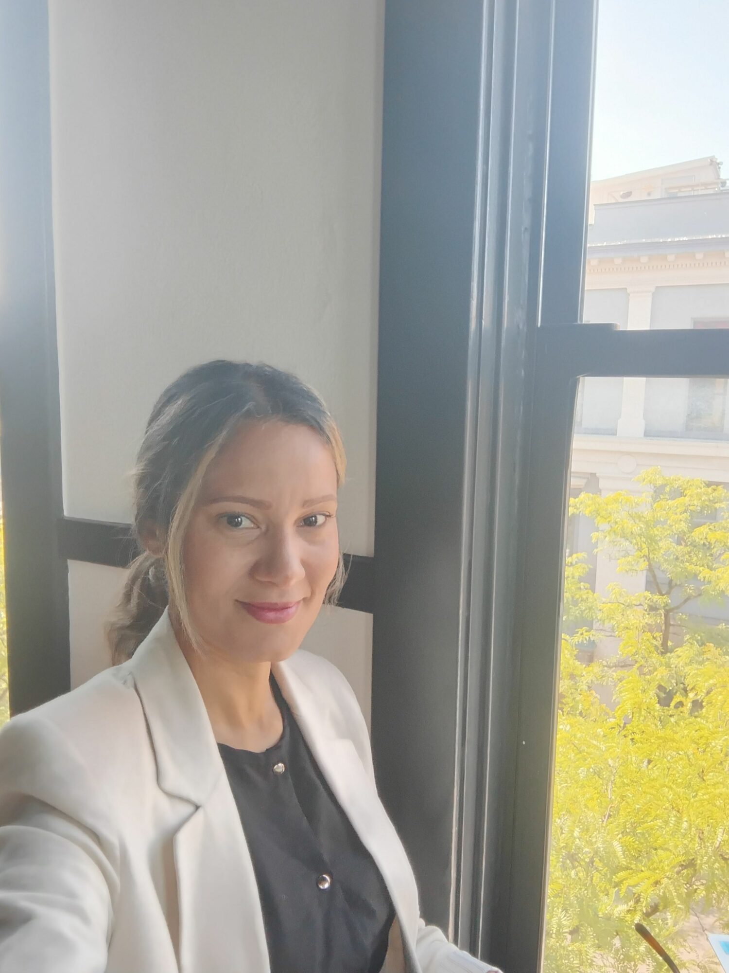 Woman with dark hair tied back, wearing a white blazer and black shirt, sitting near a window with yellow flowers outside.