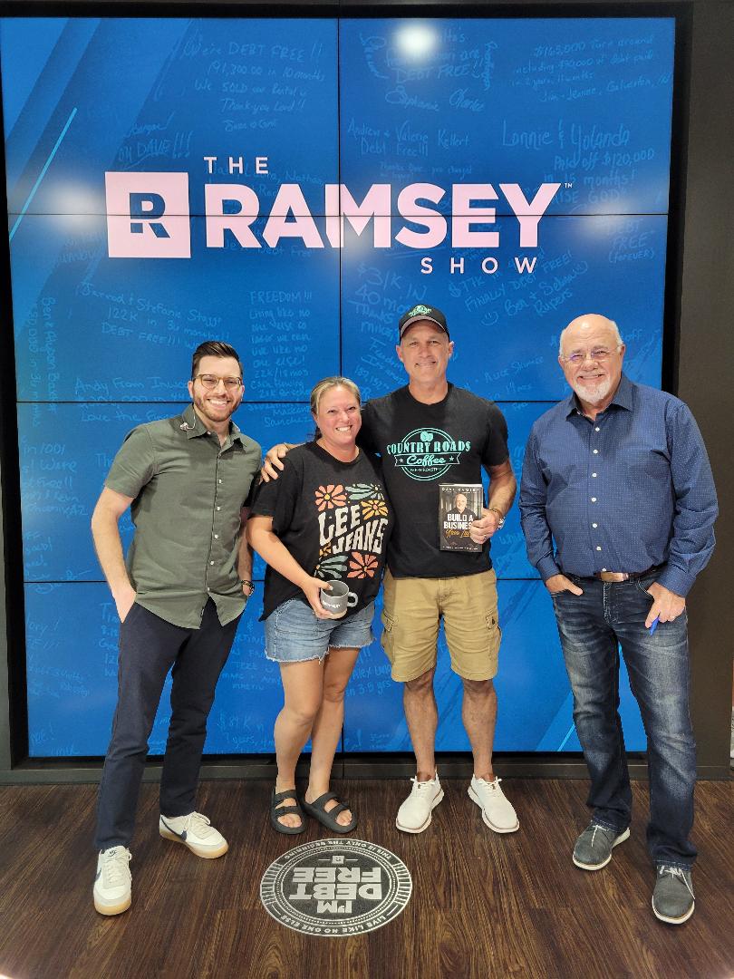 Four people standing in front of a large screen displaying 'The Ramsey Show' logo, smiling, with a wooden floor.