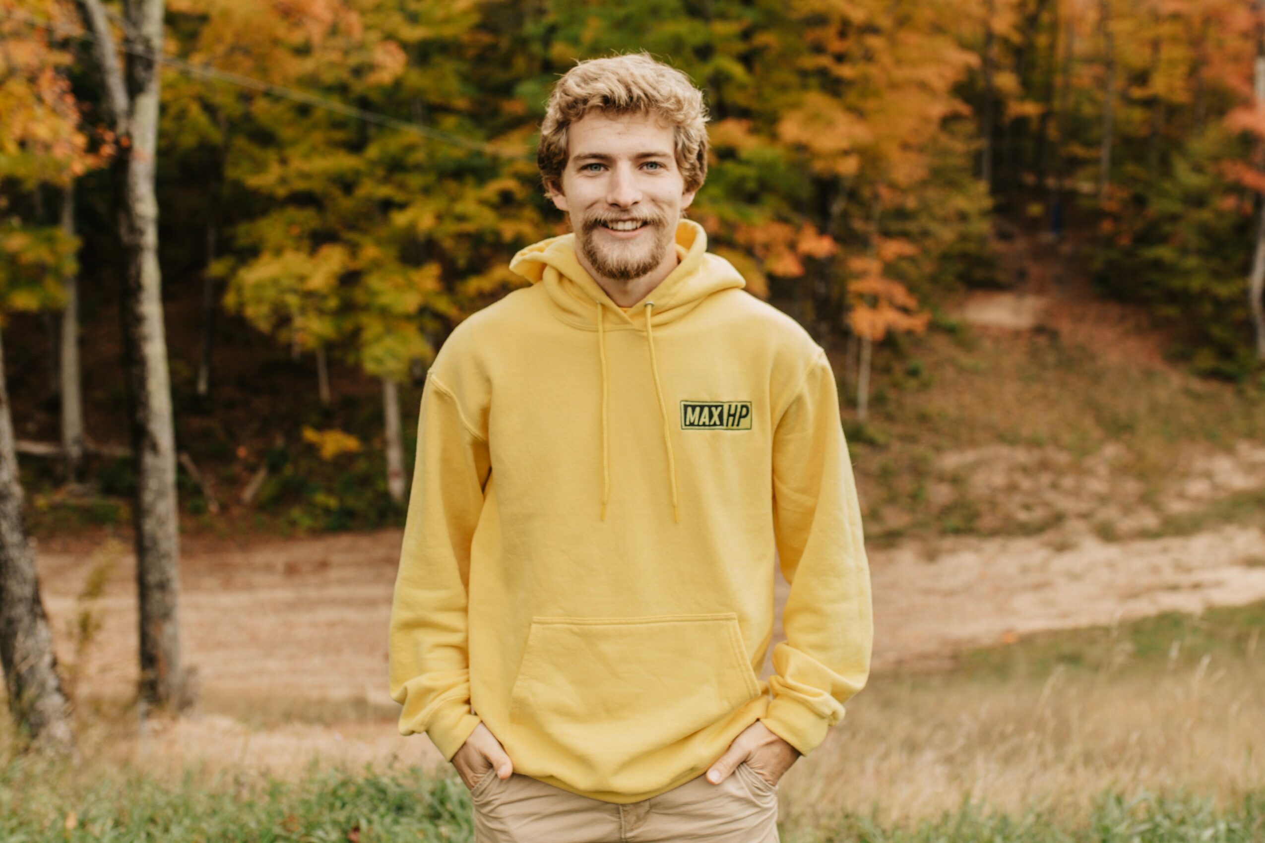 Young man with curly hair and beard smiling outdoors in autumn, wearing a yellow hoodie with a small logo, hands in pockets.