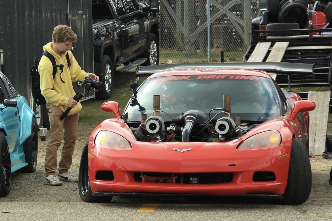 Red sports car with two large air intakes on the hood, parked outdoors with a person taking photos nearby.