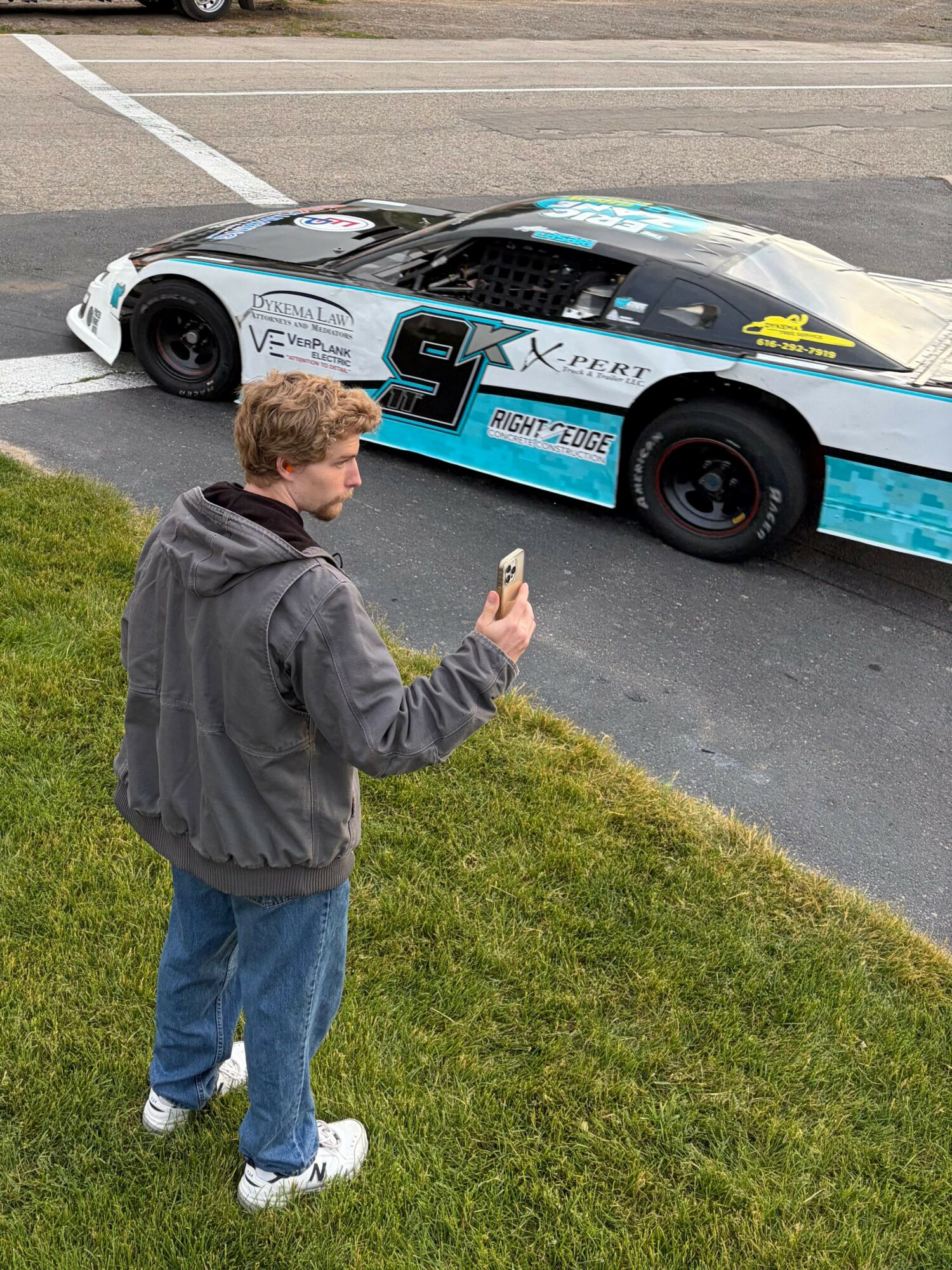 Young man in gray hoodie and jeans stands on grass, looking at a race car on the road, holding a phone.