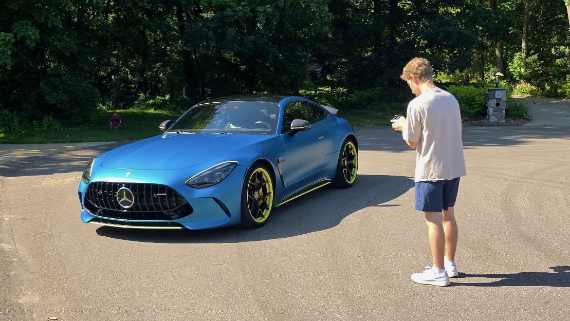 Person standing near a blue sports car on a paved road, trees in the background, sunny day.